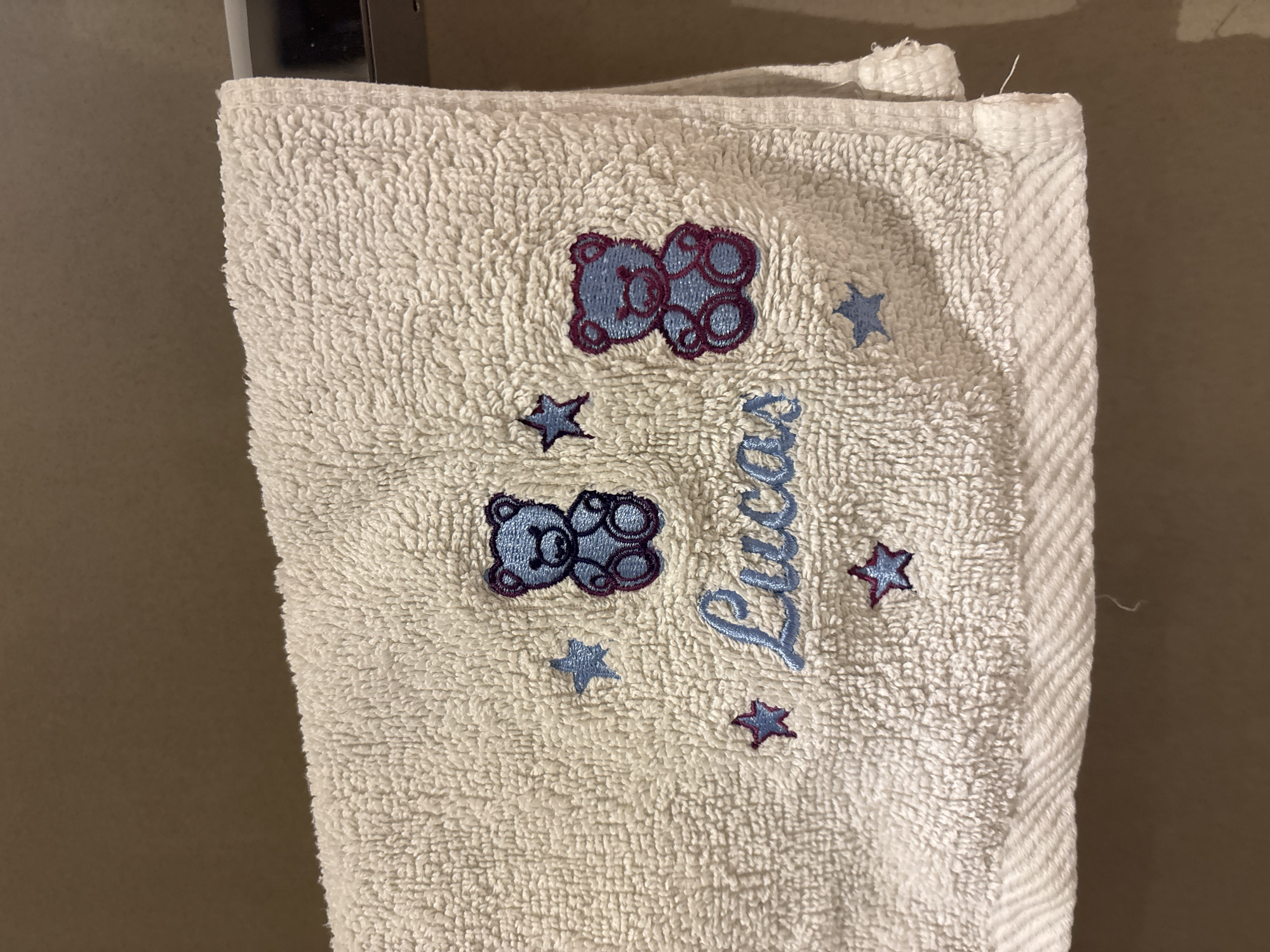 The image shows a white towel with blue and purple embroidery of two teddy bears, stars, and the name "Lucas". The towel is hanging on a silver bar against a brown, textured wall. The lighting suggests an indoor setting. There are no people or animals visible in the image. The scene does not provide any specific information about the location within Dubai, United Arab Emirates.
