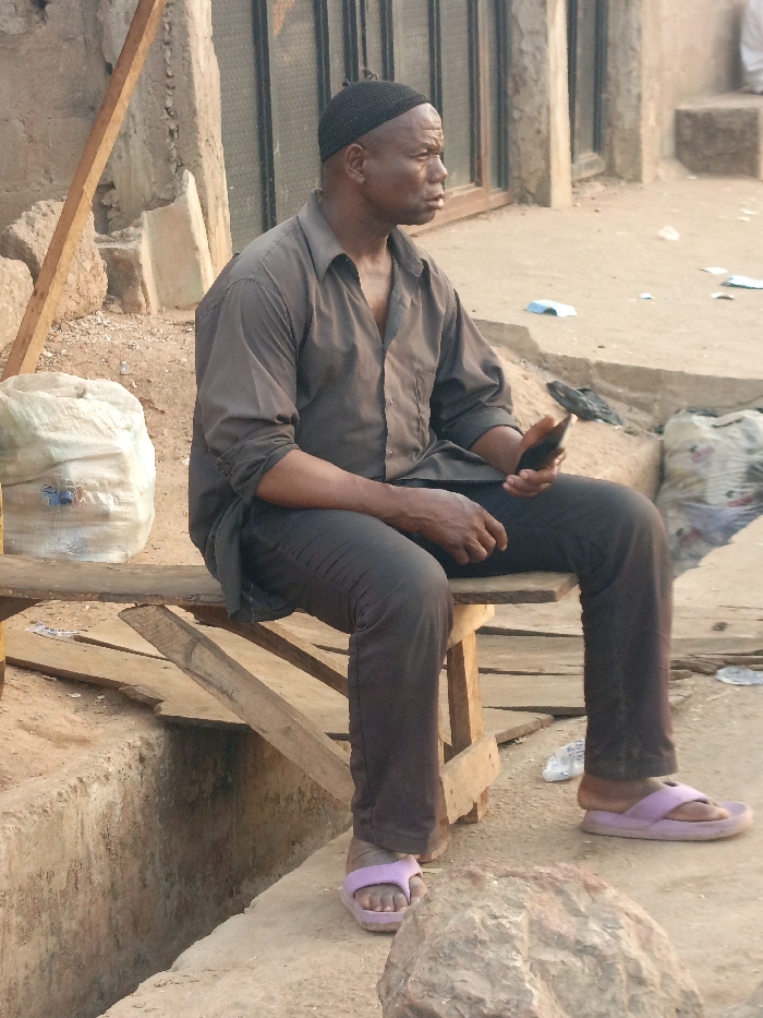 An adult male is seated on a rudimentary wooden bench in Alizaga, Nigeria. He is dark-skinned, wearing a black knitted cap, a dark grey long-sleeved collared shirt, dark trousers, and light purple flip-flops. His right hand holds a dark-colored smartphone. He faces towards the viewer's left. The environment consists of a dirt ground with scattered debris, including paper and plastic waste. To the male's left, a large white sack rests on the ground. To his right, another filled white sack is visible amidst further debris. In the immediate foreground, a light-colored rock or concrete lump is present. A concrete-lined drainage ditch runs along the seated area. In the background, unpainted or mud-colored structures with a dark-framed window or door are visible. A wooden beam leans against a wall to the left.