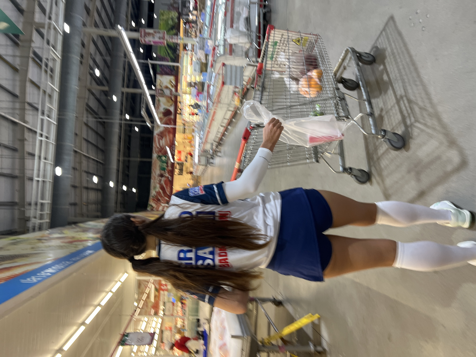 The image shows a woman with long brown hair in two braids walking away from the camera in a supermarket aisle in Baan Bo Phut, Thailand. She is wearing a white and blue sports jersey with "GRAD SA BADM" visible on the back, blue athletic shorts, white knee-high socks, and white athletic shoes. She is holding a plastic bag with her right hand and pushing a shopping cart with her left hand. The shopping cart contains some produce, including oranges and what appears to be grapes, as well as other grocery items. The supermarket is well-lit with fluorescent lighting and has multiple aisles stocked with products. Other shoppers and shopping carts are visible in the background, along with refrigerated display cases.