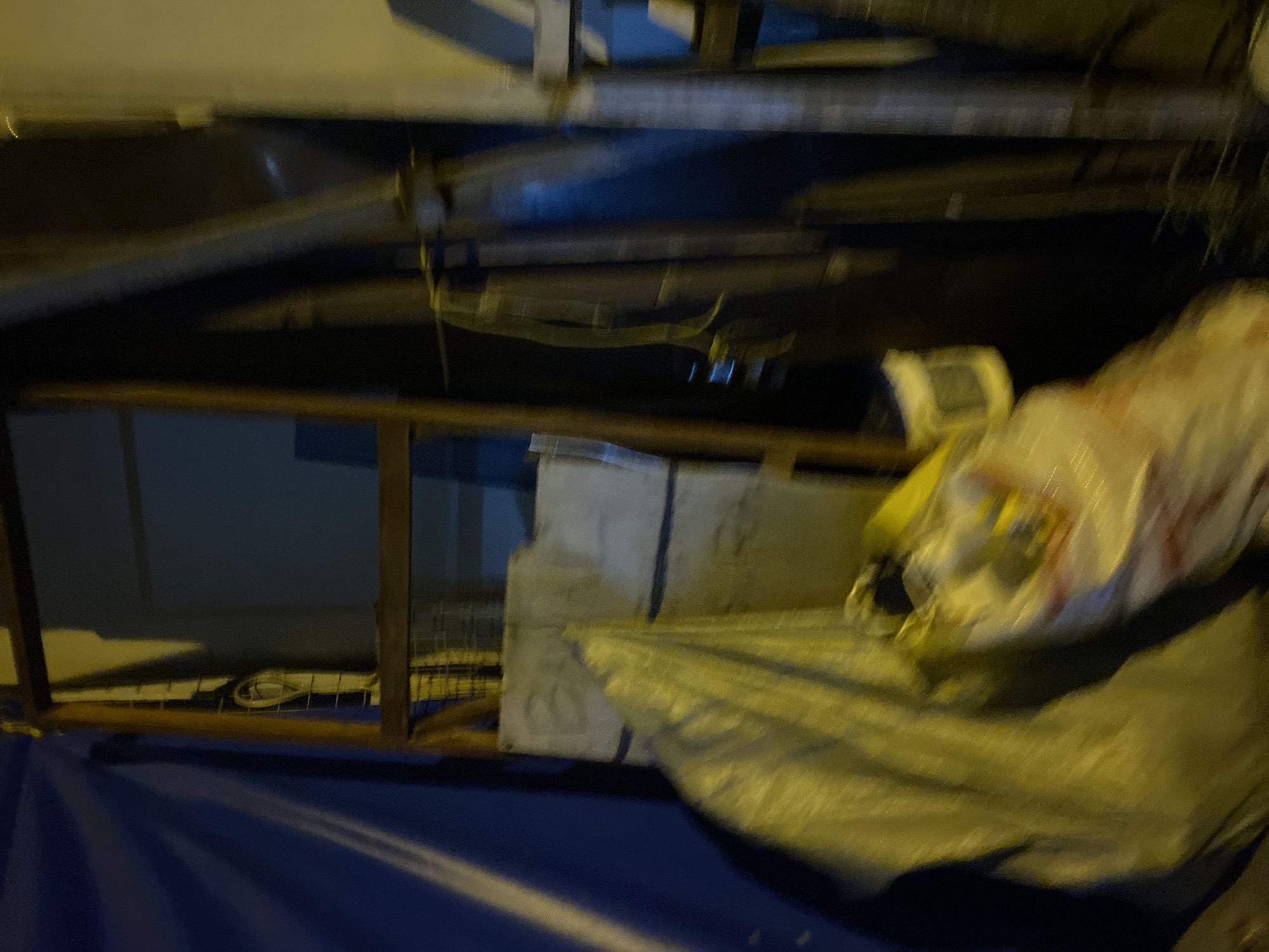 This image, captured in Davao City, Philippines, portrays a dimly lit, cluttered outdoor or semi-sheltered environment at night, characterized by significant motion blur. On the far left, a blue tarp hangs, showing prominent folds and creases, with diagonal streaks of light from an unseen source. A rough wooden frame, possibly part of a temporary structure or stall, stands centrally, extending vertically through the image. Behind the upper-left section of this frame, a looped white cable or wire is visible against a darker background. In the foreground, a large, crumpled yellow tarp or plastic sheet covers indistinct objects, intensely illuminated by a yellow light source from the left. Adjacent to this, a white bag with red markings is partially visible. Various dark, indistinct objects, including what appears to be more structural elements and possibly another blue object, are present in the background to the right of the central frame. No individuals, specific actions, or interactions are discernible.