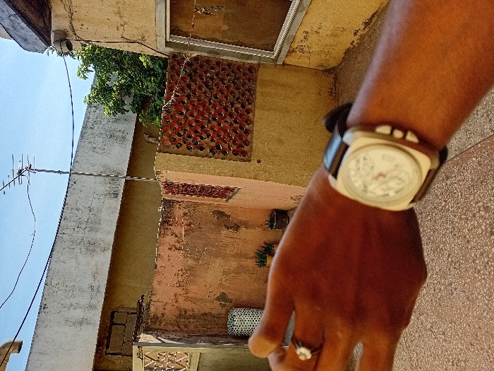 The scene, set in Sharada, Nigeria, depicts an outdoor environment under a clear blue sky. In the immediate foreground, a dark-skinned human left forearm and hand are prominently featured, presenting a first-person perspective. A square-faced wristwatch with a light-colored dial, a dark leather strap, and three visible push-buttons on the right side of the case is worn on the wrist. A silver-toned ring with a white stone is present on the ring finger. The ground surface directly beneath the hand is a light-colored, mottled concrete.
In the midground, an aged, light orange-pink plaster wall exhibiting weathering and dark stains extends horizontally. Sections of this wall are integrated with reddish-brown, circular-perforated brickwork acting as decorative screens. To the left of this wall, a white or light-colored cylindrical object with a patterned surface is visible. Small potted plants are discernible on the ground behind the orange wall. To the right, a section of a light yellow-brown building wall is visible, featuring a rectangular window frame.
The background shows the weathered, flat concrete roofline of a building. A tall metallic pole supporting a Yagi-Uda television antenna extends upward from behind this structure, silhouetted against the clear blue sky. Green tree foliage is visible to the right of the antenna. Dark-colored utility lines traverse the upper portion of the sky.