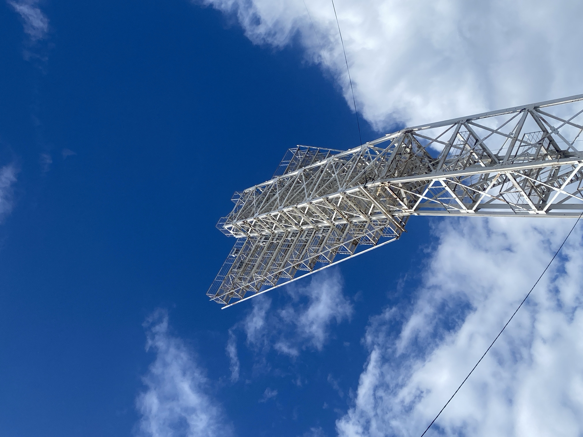 This media file captures a low-angle view of a large, metal lattice structure against a bright blue sky with scattered white clouds. The structure, which appears to be a lighting rig or part of a stadium grandstand, is made of white metal beams forming a complex geometric pattern. The perspective emphasizes the scale and height of the object, with the ground not visible. A thin black wire stretches diagonally across the frame.
The sky is a deep, vibrant blue, suggesting a clear day, possibly late morning or early afternoon, given the brightness. The clouds are wispy and white, indicating fair weather. There are no people visible in the frame. The overall impression is one of architectural grandeur and an expansive, open environment. The location context of Заволжье, Russia, is noted, but no specific geographical landmarks are identifiable within the image itself. The image is devoid of any visible text. The scene is neutral in emotional tone, focusing on the visual details of the structure and the sky.