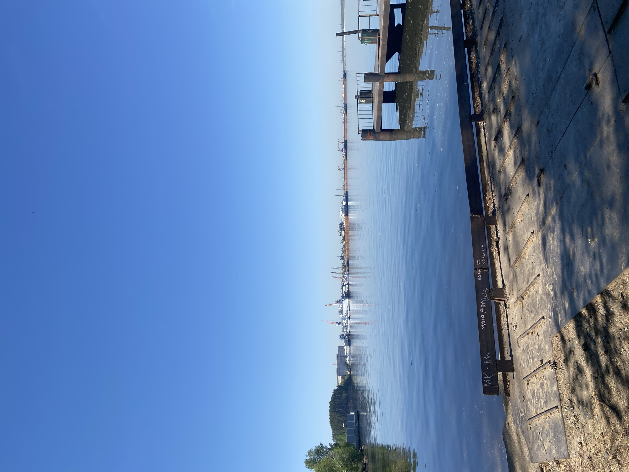 The image shows a waterfront scene in Chaikovsky, Russia. The sky is clear and blue. Calm water fills the majority of the frame, reflecting the sky and distant structures. A dilapidated concrete pier extends into the water on the right side of the image. Graffiti is visible on the side of the pier. In the distance, there are industrial buildings and cranes. Green trees are visible at the water's edge in the lower center of the image.