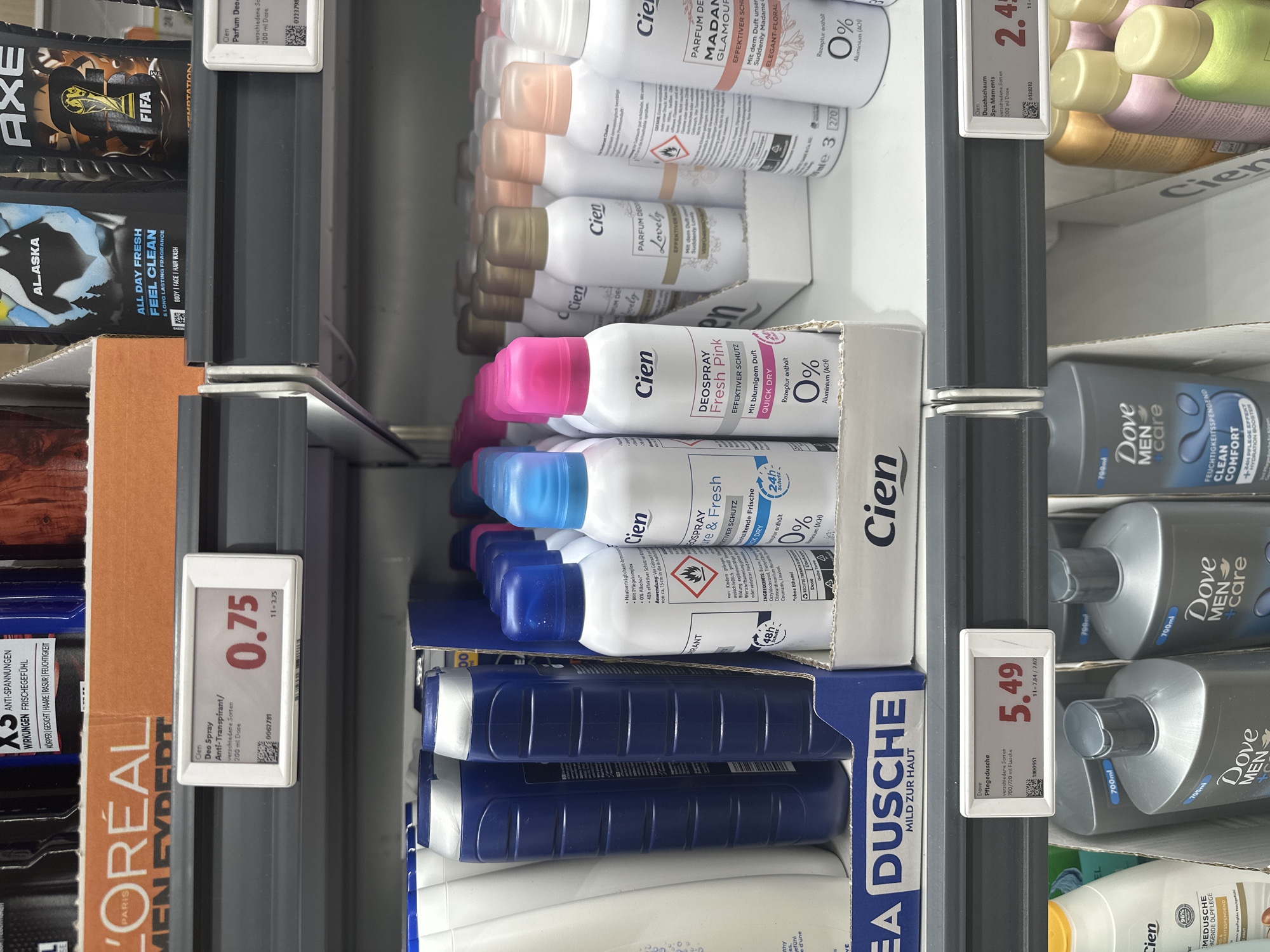 The image shows a supermarket shelf displaying various personal care products. In the foreground, there are several Cien brand deodorant sprays with colorful caps, and Dove Men+Care body washes. Further back, larger bottles of what appear to be Cien shower gels are visible. A shelf above holds bottles of L'Oréal Men Expert products and other deodorants from brands like Axe. Price tags are visible on some of the products. The lighting suggests an indoor retail environment. The overall scene is a typical retail display of toiletries. This image was taken in a supermarket in Freiburg im Breisgau, Germany.