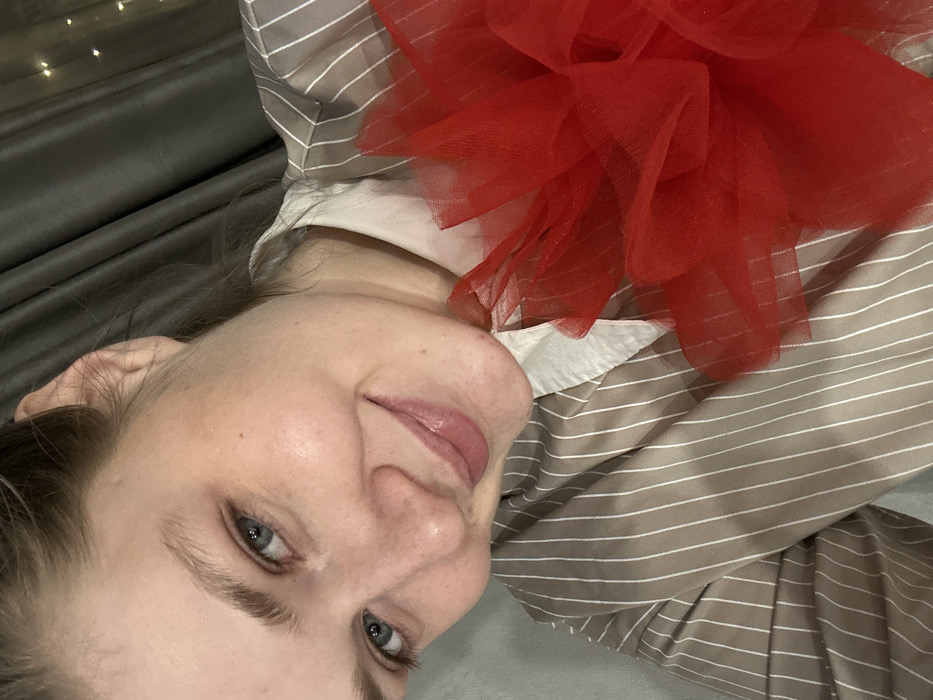 The image displays a female subject indoors in Borovoye Matyushino, Russia. The subject, an adult with light skin, blue eyes, and light brown eyebrows, is depicted from the upper torso upwards, angled slightly towards the camera. Her hair is pulled back, exposing her right ear. She is smiling, looking directly at the viewer, with slightly parted lips enhanced with a light pink gloss. She wears a light brown or taupe button-up shirt featuring thin white vertical stripes, with a white collar visible beneath. A substantial, multi-layered object constructed from bright red tulle or similar sheer fabric rests on her right shoulder and chest area. The immediate background consists of a solid light gray wall or surface on the left and dark gray curtains on the right. Blurred, faint lights, consistent with string lights, are discernible through or behind the curtains on the upper right side of the frame.