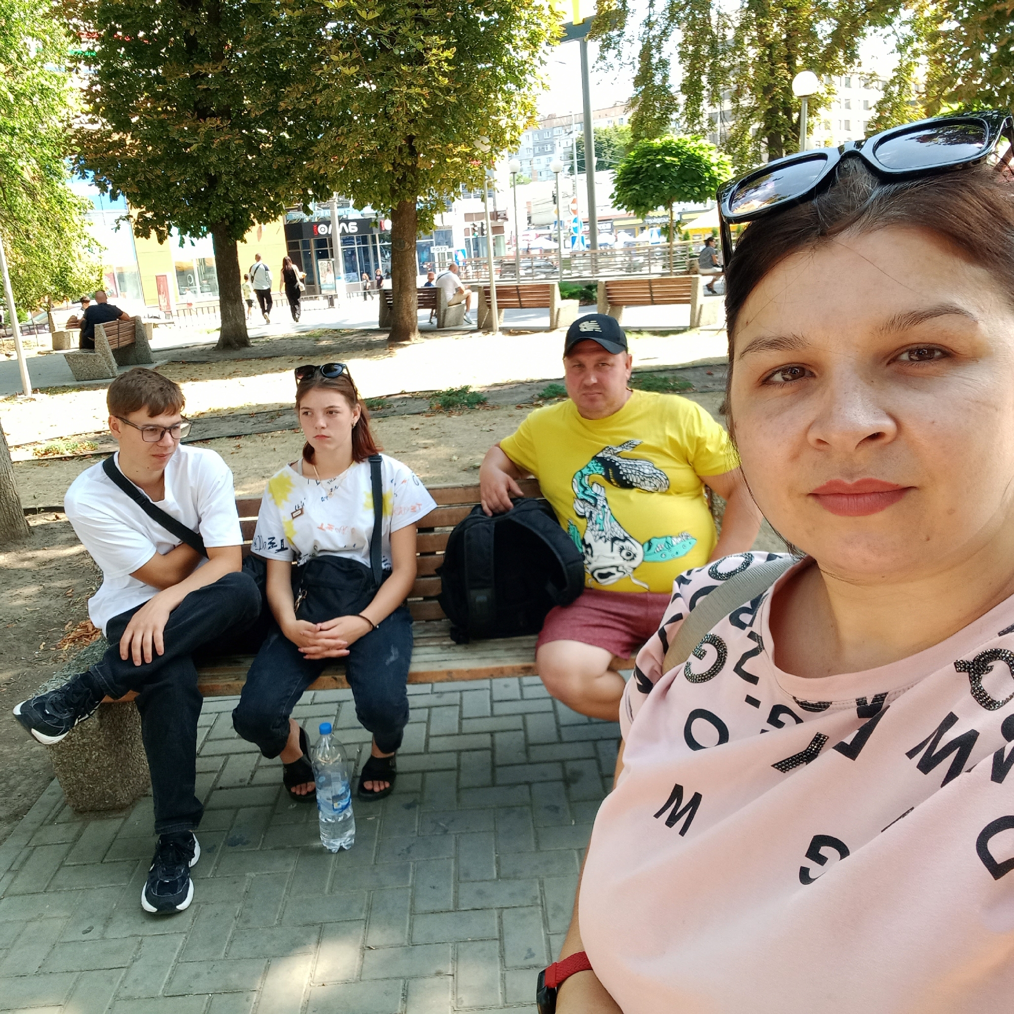 The image depicts a group of people sitting on a park bench in Kropyvnytskyi, Ukraine. The group consists of two young adults, a man, and a woman. The young adult male, wearing glasses and a white t-shirt, sits on the left side of the bench with his arms crossed. Next to him is a young adult female in a white t-shirt with yellow markings, looking to her left. A man in a yellow t-shirt and a black baseball cap is seated to the right of the young woman, with a backpack beside him. The woman taking the photo is in the foreground, with a pink t-shirt adorned with black letters and sunglasses on her head. A bottle of water is placed on the ground next to the bench. The background shows trees, a street with buildings, and lampposts, suggesting an urban park setting within the city.