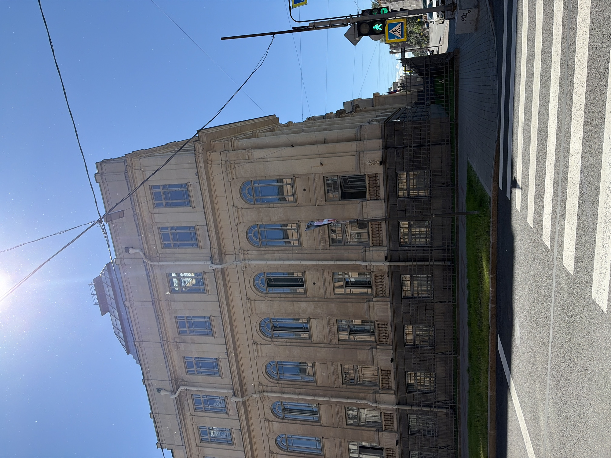 The image captures a sunny day in Saint Petersburg, Russia, focusing on a grand, light-colored building with classical architectural elements. The building features multiple stories with arched windows on the upper levels and rectangular windows below. A dark, ornate fence runs along the base of the building, separating it from a strip of green grass and then a paved sidewalk. In the foreground, a pedestrian crossing with white stripes is visible on the asphalt road. To the right of the building, a traffic light displaying a green pedestrian signal and a yellow pedestrian crossing sign are mounted on a pole. Overhead, power lines are strung across the clear blue sky, and the sun is shining brightly, causing some lens flare. The overall scene depicts a typical urban streetscape in the city.