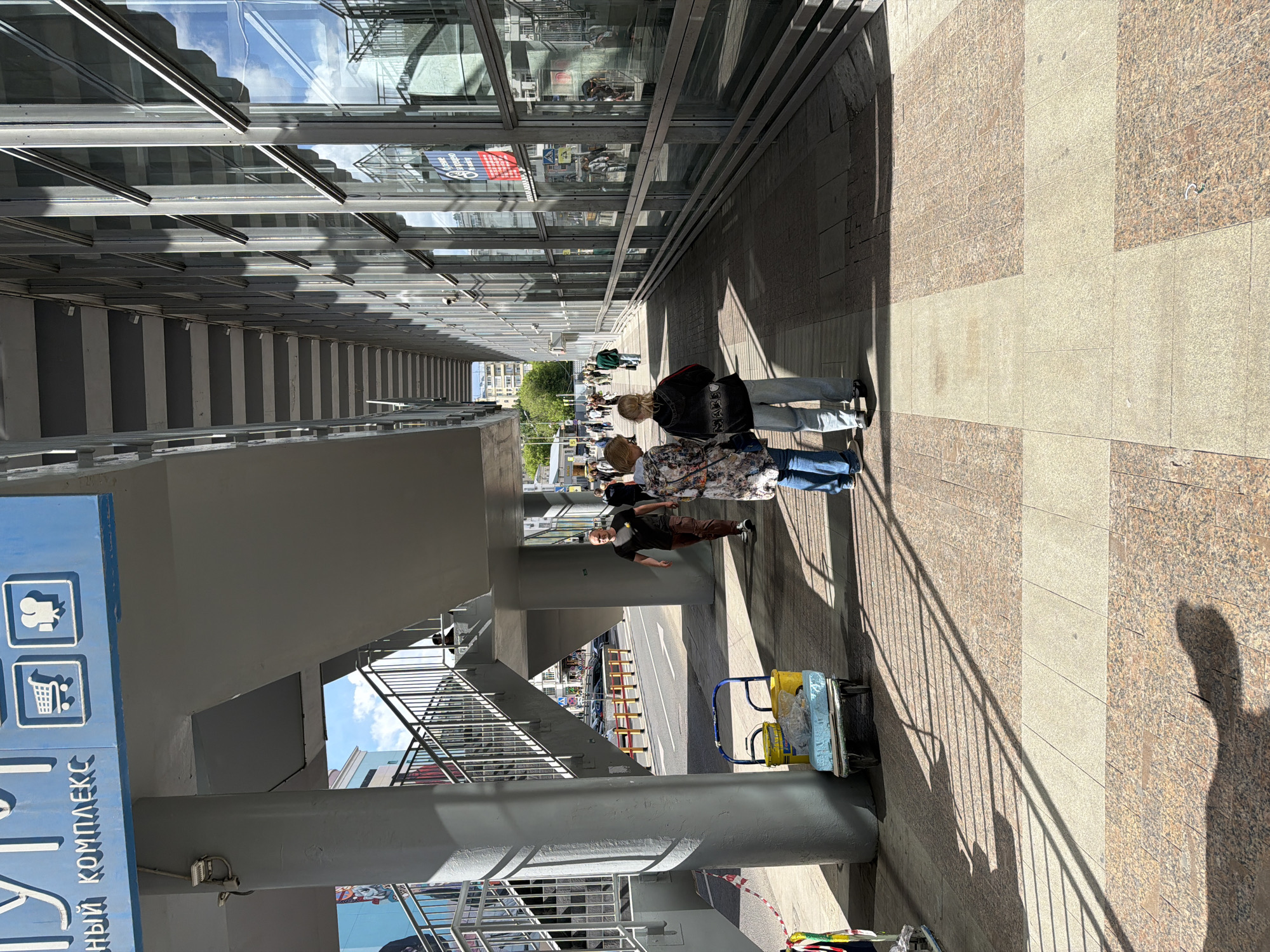 Here is a detailed description of the image:
The image captures a public space in a city, potentially in an area with commercial activity, as indicated by signage on the left side of the frame. The scene is oriented at an oblique angle, creating a sense of perspective along a walkway or sidewalk.
Along the walkway, a small group of people is present. Two women are walking side-by-side toward the end of the walkway. Beside the end of the walkway there is a man standing with a cart beside him. There is a man walking between the two women and the man with the cart. The sidewalk is paved with a patterned stone surface, with alternating bands of lighter and darker stone.
The walkway is partially covered by an overpass-like structure with supports that cast shadows on the pavement. To the left of the walkway, there is a blue sign displaying symbols and text, potentially indicating services or facilities in the area.
Above the walkway, there is a section of a building with numerous windows, reflecting the sky. The overall environment suggests an urban setting with a combination of architectural elements, pedestrian activity, and signage.