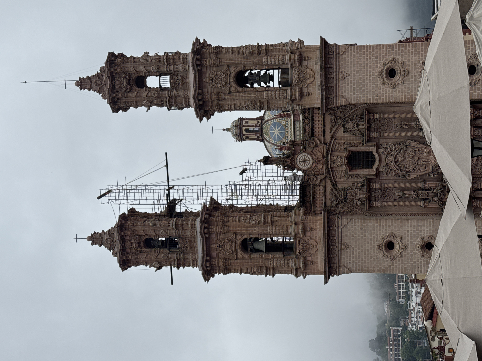 The image shows a close-up of the ornate facade of the Santa Prisca Church in Taxco, Mexico. The church, constructed from pink stone, features two prominent bell towers with intricate carvings and crosses at their apex. Scaffolding is visible around the upper sections of the towers, suggesting ongoing restoration or construction work. A colorful tiled dome is positioned between the towers, topped with a smaller dome and a cross. Below the dome, a clock face is visible on the facade, along with detailed sculptural elements and windows. The lower portion of the image shows the tops of several large, light-colored umbrellas, obscuring the view of the street level. In the background, a misty hillside is dotted with white buildings and lush greenery, indicating a mountainous and somewhat overcast environment. The overall impression is of a grand, historic religious building in a potentially humid or foggy climate.