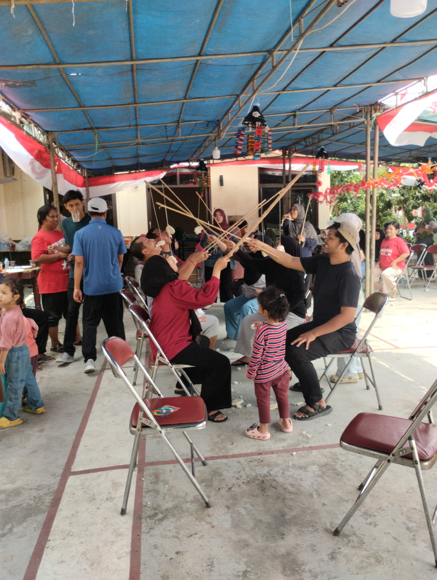 This image depicts an outdoor gathering under a blue tarp roof supported by a metal frame, likely in Bekasi, Indonesia, given the red and white Indonesian flags visible. Several people are participating in a game where they are using long wooden sticks to try and capture food items suspended from above. In the foreground, a young child wearing a pink and red striped shirt and pink sandals stands on the concrete ground. To the right, a man in a black shirt and a beige cap is seated on a red folding chair, holding a stick. Other participants, including women and men, are seated or standing, engaged in the activity. Scattered pieces of white food, possibly crackers or snacks, are visible on the ground. The environment appears to be a communal area, possibly a yard or a public space, with concrete flooring and some vegetation in the background.