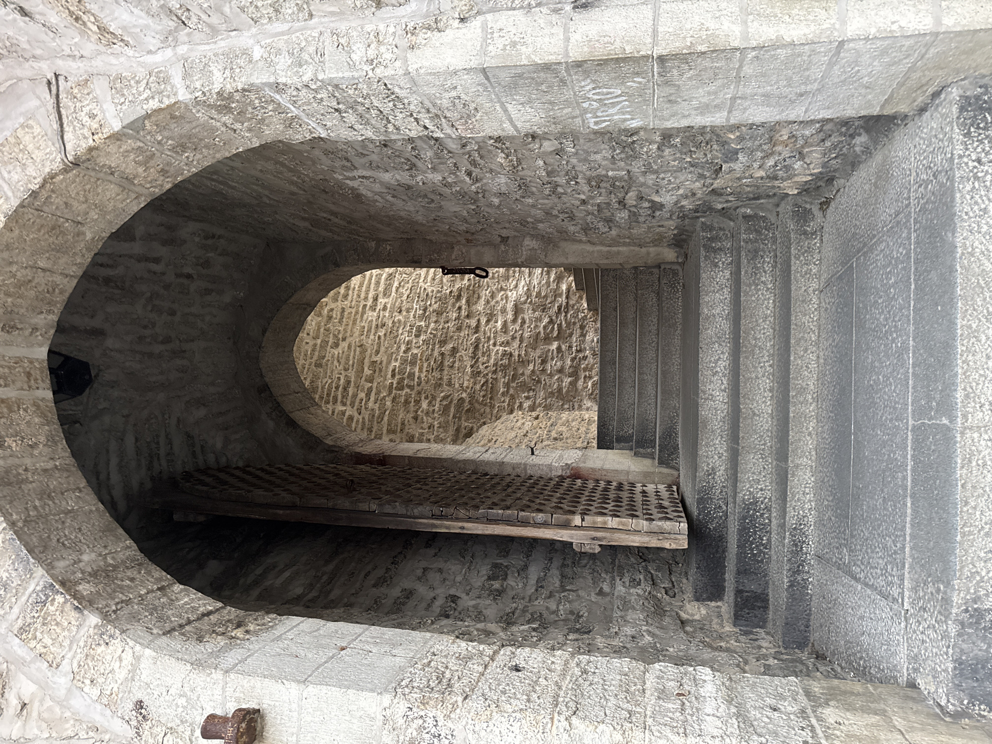 The image shows a stone archway leading to a set of stone stairs. The archway is made of rough-hewn stone blocks. A large wooden gate with metal studs is partially open to the left of the stairs. The stairs are made of dark gray stone and ascend into a narrower, arched passageway. The walls are made of stone, and there is graffiti on the right-hand wall. This is likely the Viru Gate in Tallinn, Estonia, which is an iconic medieval city gate and part of the old town walls.