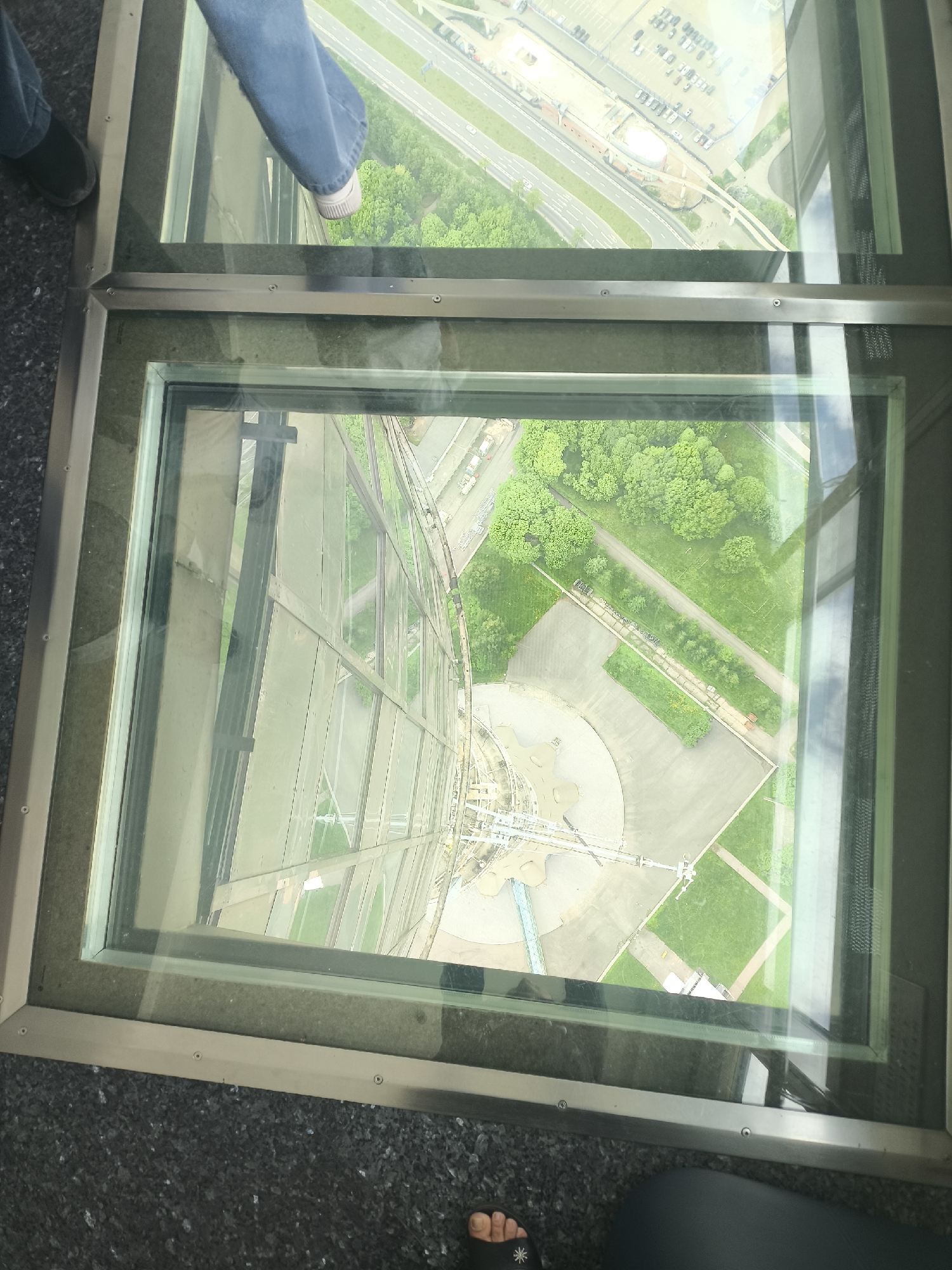 The image shows a view from a high vantage point through a glass floor or panel, possibly in a skyscraper or observation tower. The location is likely Khimki, Russia, as inferred from the landscape seen through the glass.
**People:** Two individuals are partially visible. One individual is wearing jeans and shoes, with their feet placed on the glass panel in the upper part of the image. The other individual is at the bottom of the image wearing sandals, with only toes visible.
**Subjects:** The primary subject is the downward view through the glass panels. The cityscape below includes buildings, trees, roads, and green spaces. The view appears to be directed towards a complex that may contain industrial structures or construction sites.
**Objects:** The prominent objects are the glass panels, which are framed by metal supports. There is a structure below that appears to be under construction and greenery visible in multiple locations.
**Actions:** The action depicted is viewing or standing on the glass panel, which offers a direct view of the scene below.
**Interactions:** There is an implicit interaction between the viewers and the environment, where they are observing the landscape from a considerable height.
**Environment:** The environment includes both the interior space of the building or tower (represented by the glass panels and metal frame) and the external cityscape of Khimki. The overall atmosphere suggests a clear day, allowing for detailed visibility of the area below.