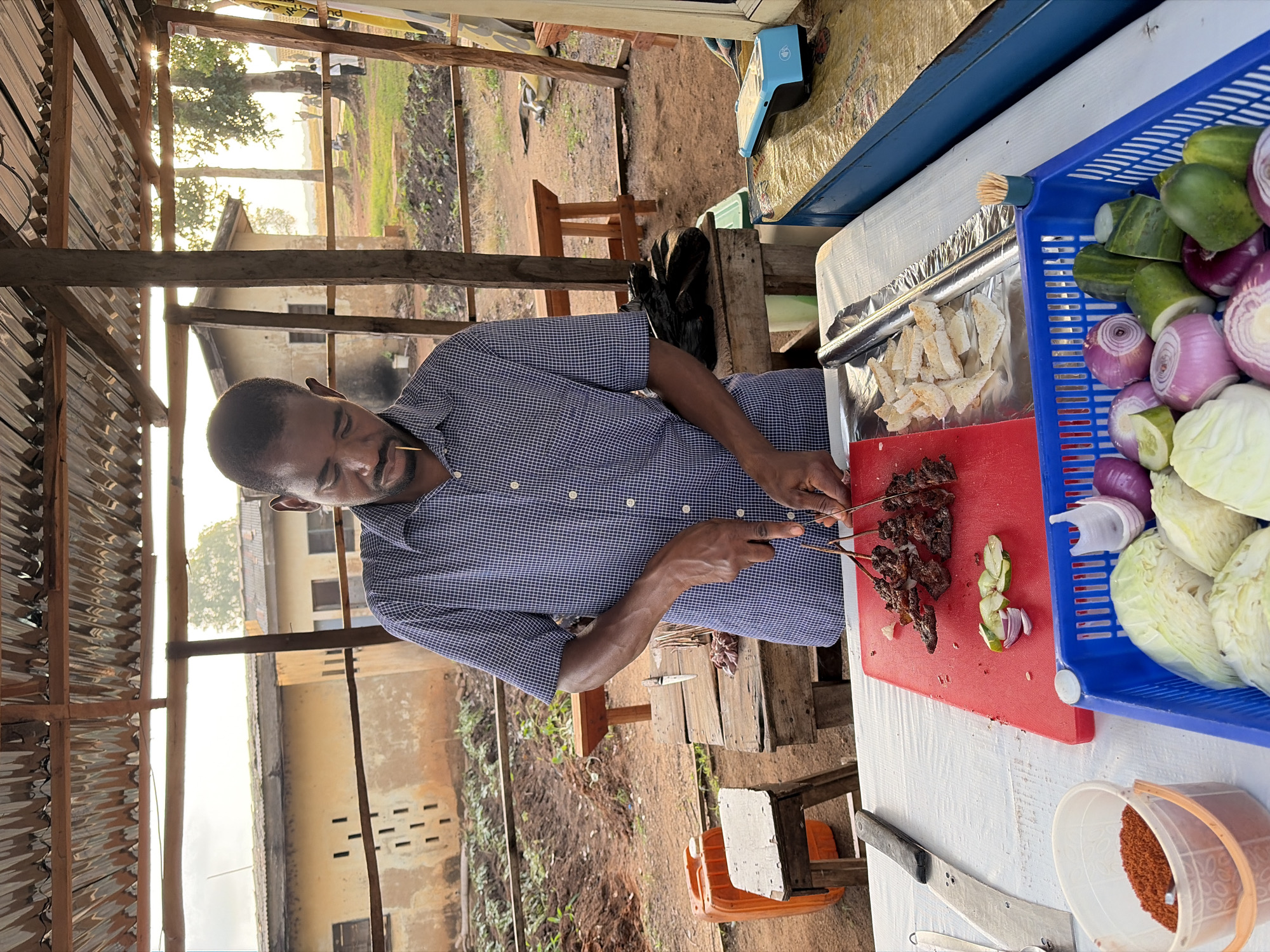 In Rafin Par, Nigeria, a man stands at a stall under a corrugated metal roof supported by wooden beams, preparing food. He is wearing a blue and white checkered shirt and is in the process of skewering meat on thin wooden sticks. In front of him, on a white tablecloth, is a red cutting board with the prepared skewers and some sliced red onion and cucumber. To the right of the cutting board, on aluminum foil, are slices of bread. A blue plastic basket containing cabbage, red onions, and cucumber slices sits on the table. To the left of the cutting board is a large knife and a white plastic container filled with a reddish-brown spice. In the background, there are several buildings with light-colored walls and dark windows, along with dirt ground, vegetation, and trees. A bright orange plastic container and a wooden stool are visible to the left of the stall. A blue electronic point-of-sale device is placed on a blue counter to the right of the stall.