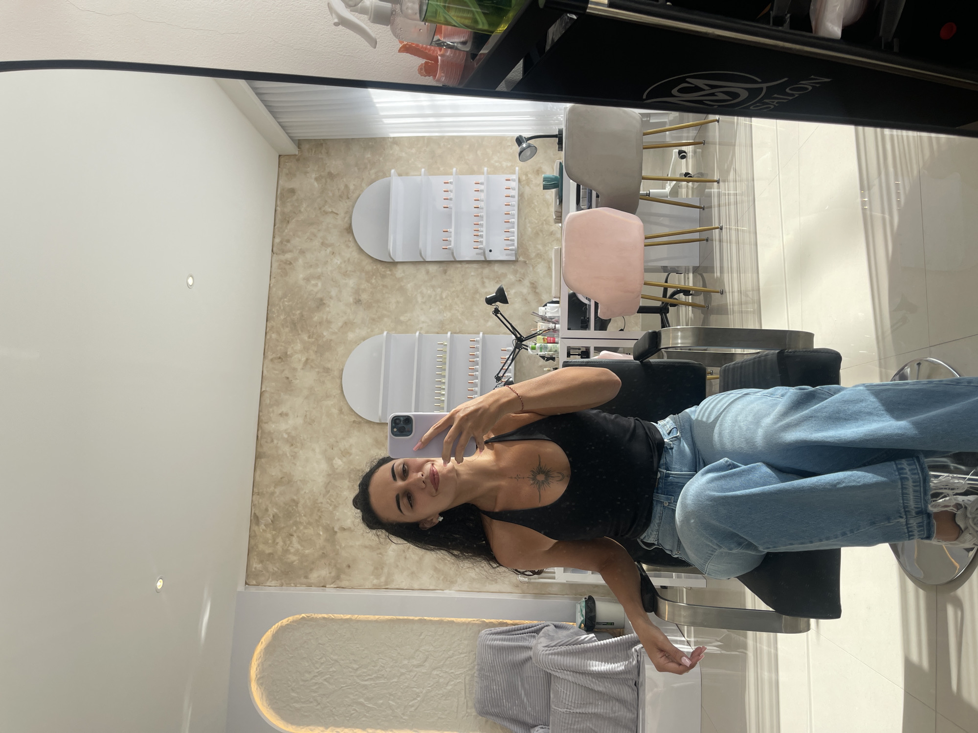 A woman is reflected in a mirror, seated in a salon-type chair with a black back and silver frame. She is wearing a black tank top and light blue jeans. She holds a smartphone up and is taking a selfie, making a peace sign with her fingers. Behind her, salon equipment and furniture are visible, including white and gray chairs, manicure tables, and wall-mounted organizers holding nail polish. The background walls are a mix of beige textured wallpaper and white tile. The salon appears modern and well-lit. The location is North Kuta, Indonesia.