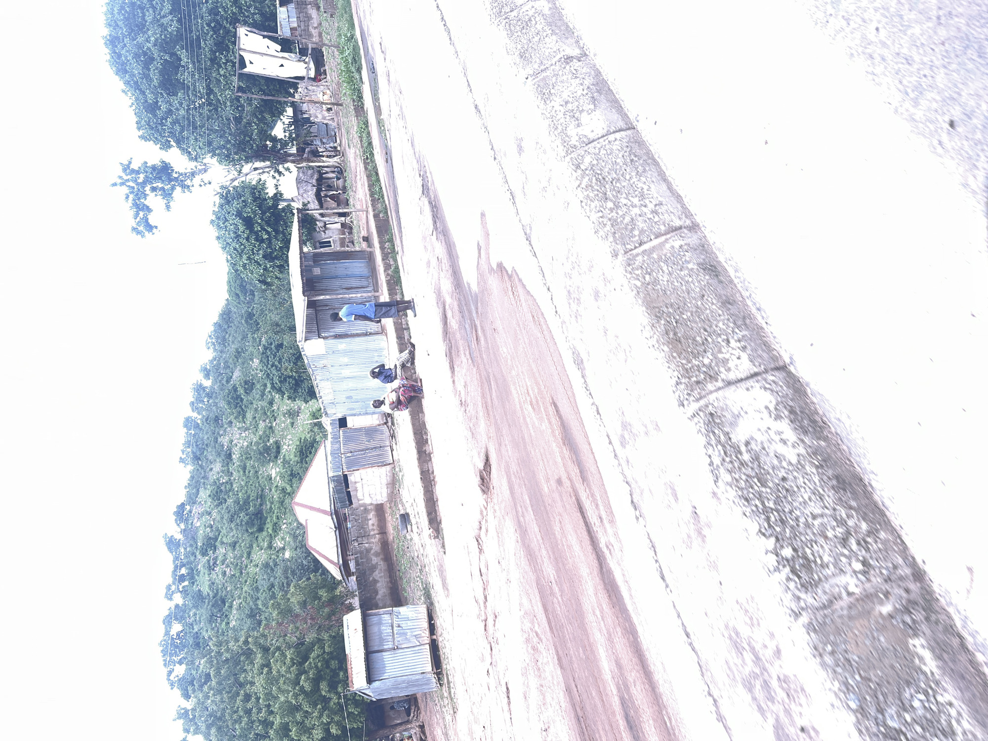 In Minda, Nigeria, a street scene unfolds with a dry, reddish-brown dirt road in the foreground, bordered by a concrete curb and gutter. Beyond the road, a collection of structures lines the path. To the left, a small, corrugated metal shed stands. Further back, several other corrugated metal buildings and a building with a red-tiled roof are visible, some appearing to be shops or residences. A few individuals are present: one person in a blue shirt and grey shorts stands near the metal shed, facing away from the camera. Two other people are seated on a low wall in front of some buildings, one wearing a patterned skirt. The background features a lush green, tree-covered hillside under a bright, overcast sky. The environment suggests a rural or semi-urban setting with simple infrastructure.
