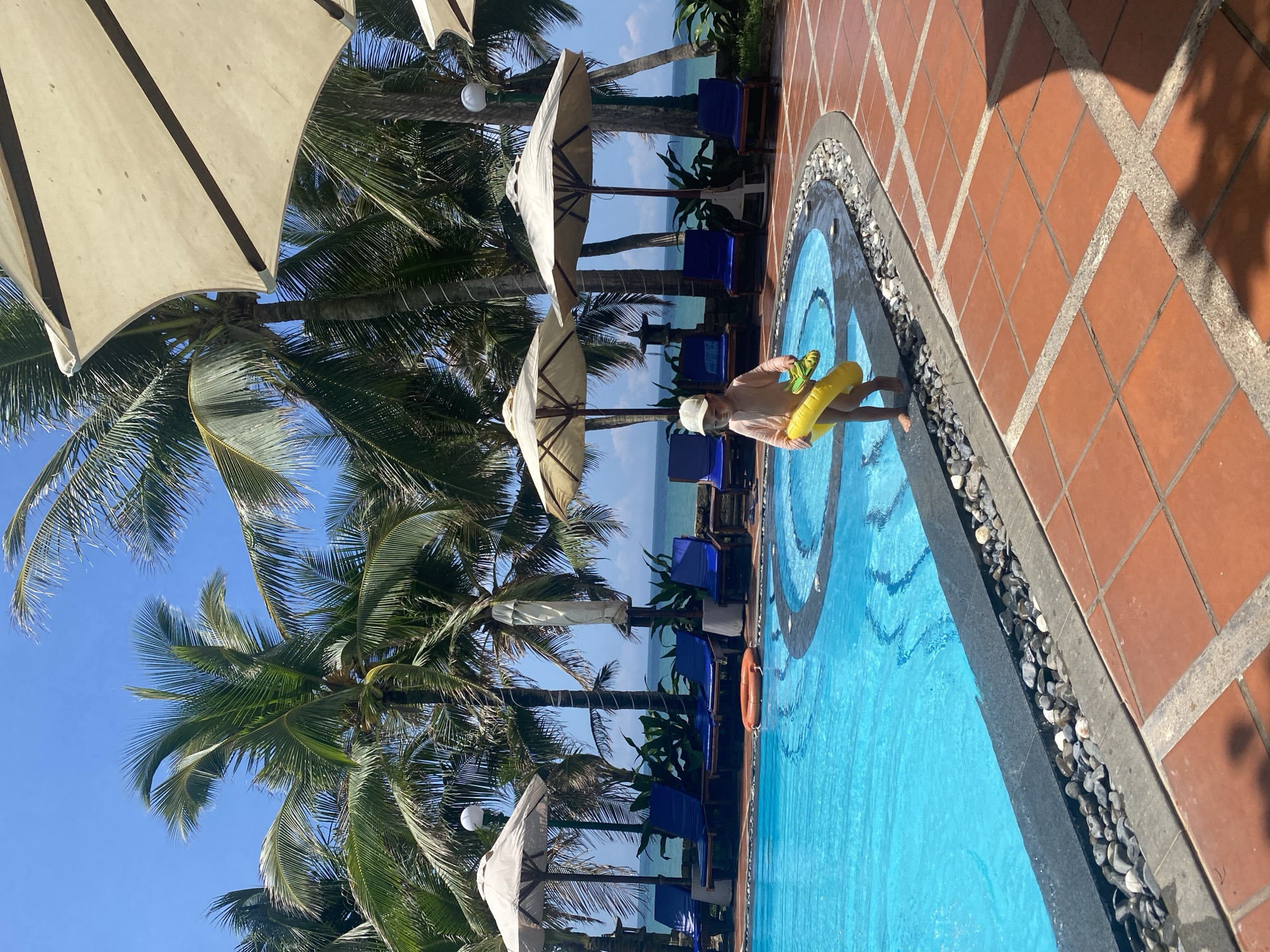 The image depicts an outdoor swimming pool area in Phu Thuy Ward, Vietnam, under a clear blue sky. A young child, wearing a light-colored long-sleeved swimsuit and a baseball cap, stands barefoot at the edge of a curved swimming pool. The child holds a deflated yellow inflatable ring in their left hand and a small yellow water toy in their right hand. The pool features light blue water and a dark grey coping, with a border of small grey and white pebbles between the coping and the surrounding terracotta-tiled patio.
In the background, multiple tall palm trees with green fronds are visible, indicating a tropical environment. Several large, off-white sun umbrellas are open, providing shade over blue lounge chairs positioned on the tiled area beyond the pool. Behind the lounge chairs and foliage, the calm, turquoise water of the ocean is visible, extending to the horizon. A single orange lifebuoy rests on the ground near the far edge of the pool. The scene is brightly lit by natural sunlight, casting shadows on the terracotta tiles.