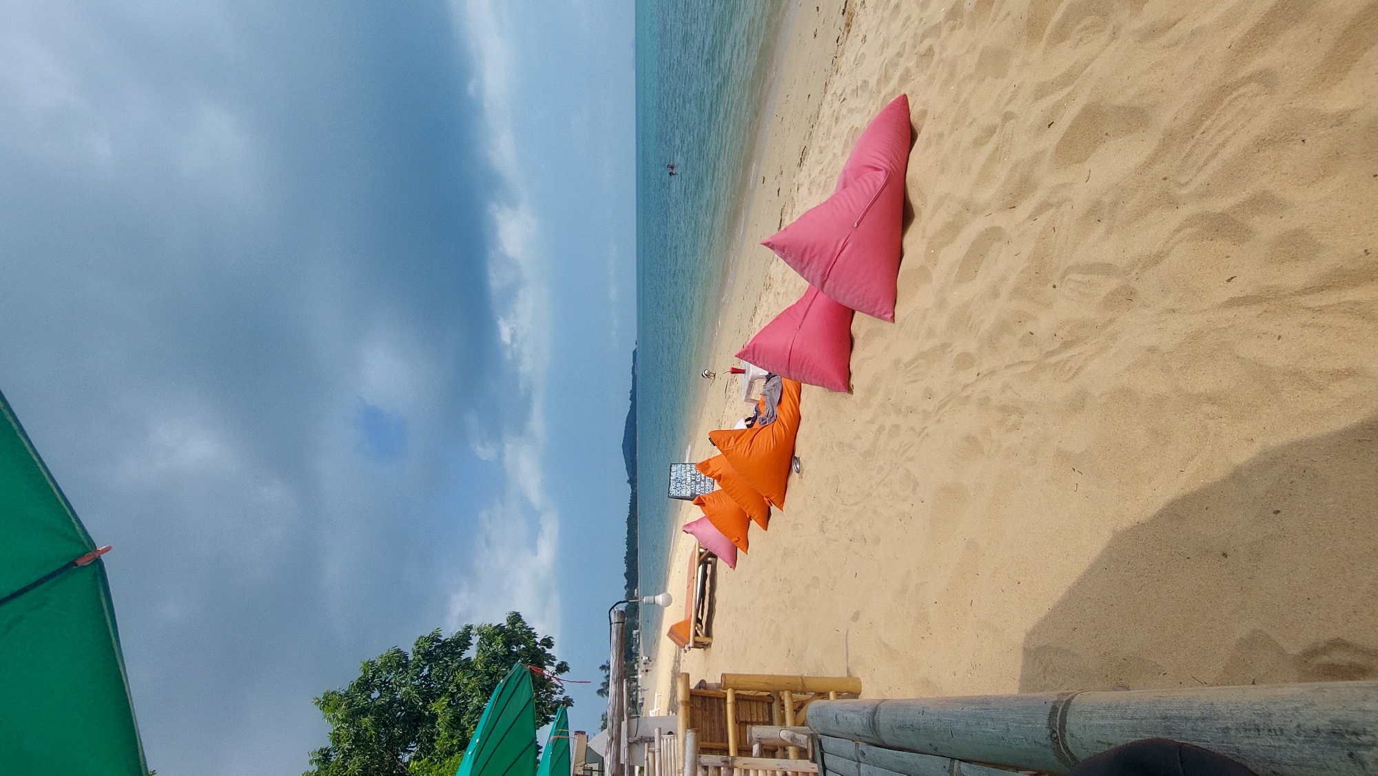 The image captures a beach scene in Baan Mae Nam, Thailand. The foreground shows bamboo furniture and green umbrellas. Beyond this, the sand is populated with a series of colorful, triangular-shaped cushions in pink and orange. In the distance, the ocean stretches to the horizon under a cloudy sky. A solitary person is swimming in the ocean.