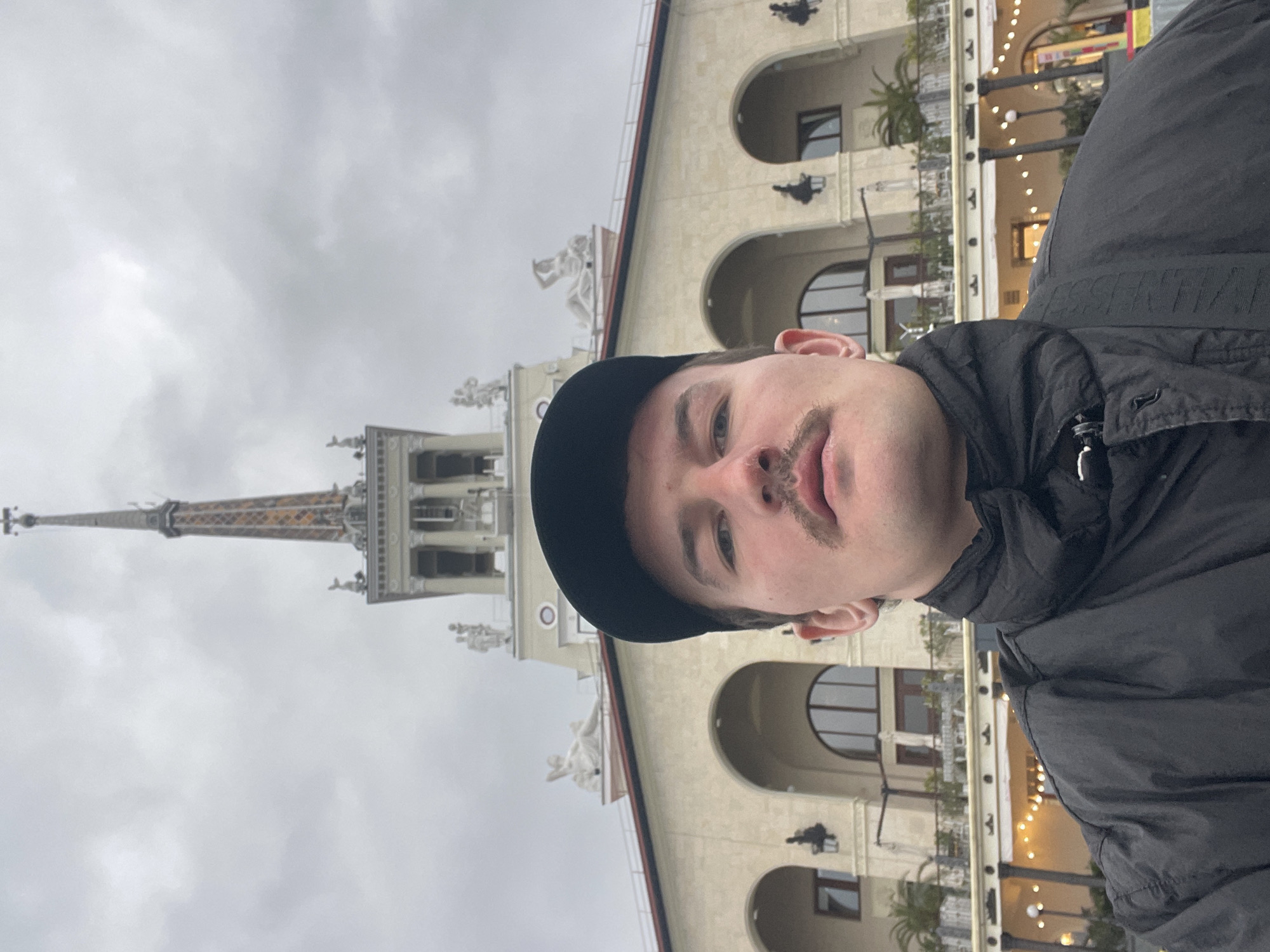 A man with a black cap and mustache is the primary subject. He's wearing a black jacket. The background is a building with a prominent spire, statues, and architectural details. This is likely the Sochi Marine Terminal in Sochi, Russia, due to the architectural style. The sky is overcast.