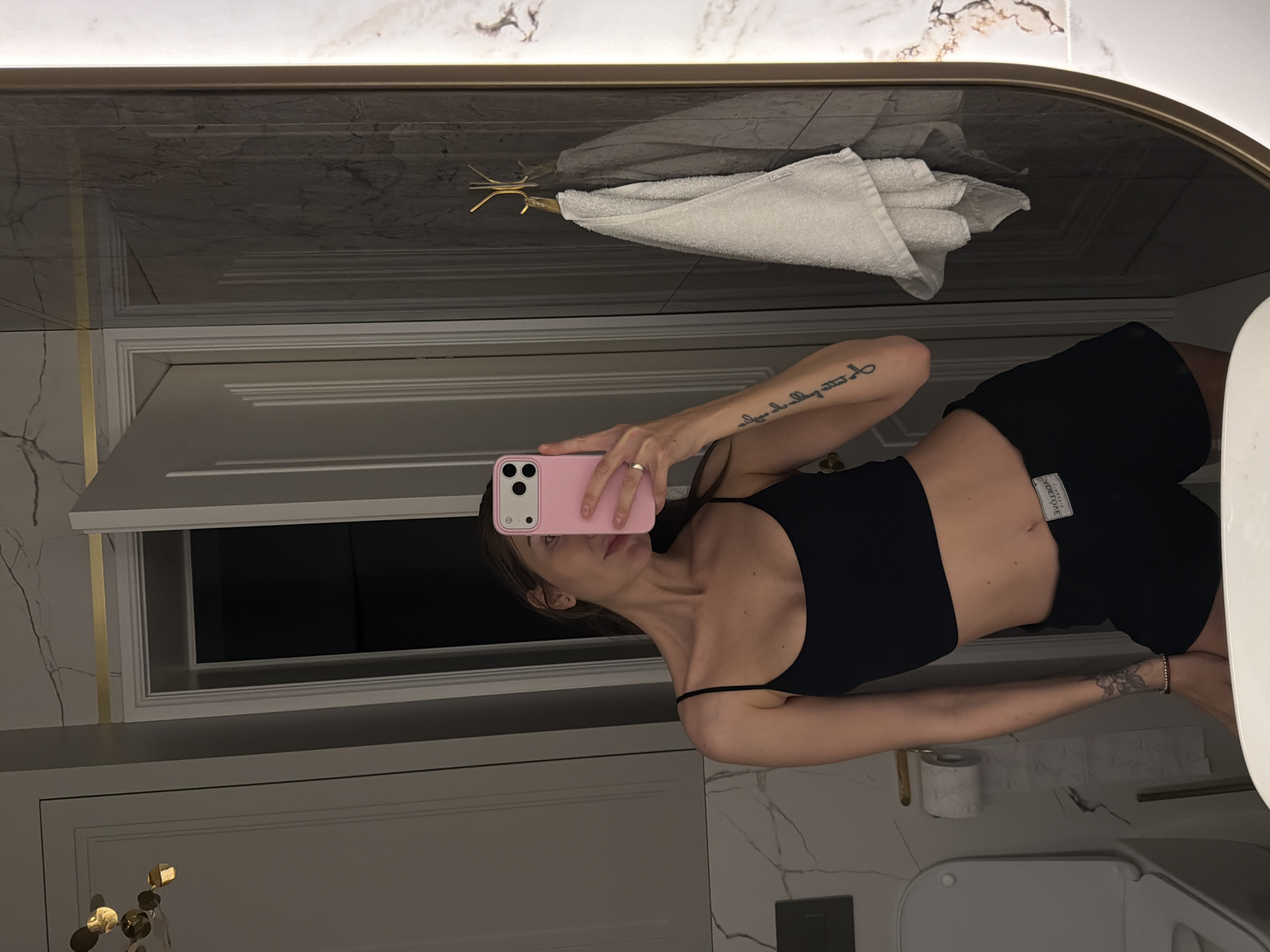 This image depicts a mirror selfie taken by a person in a modern bathroom, likely in Krasnoyarsk, Russia. The person, mostly visible from the chest down, holds a pink smartphone with a triple camera array, obscuring part of their face. They are wearing a black spaghetti-strap crop top and black shorts with a white label featuring text, possibly "ADEONE" or similar, on the right hip. A faint scar is visible on their lower abdomen.
The person has a script tattoo on their left forearm and a floral or abstract design on their right wrist/forearm, and wears a ring on their right hand. The bathroom exhibits a luxurious design with white and grey marble-patterned walls and surfaces. A large mirror with a golden frame reflects a folded white towel hanging from a golden, multi-pronged fixture above white paneled cabinetry or doors accented with golden trim. A toilet paper roll on a golden holder and a partial view of a white toilet bowl are visible in the reflection. The lighting is artificial, illuminating the sophisticated interior.
