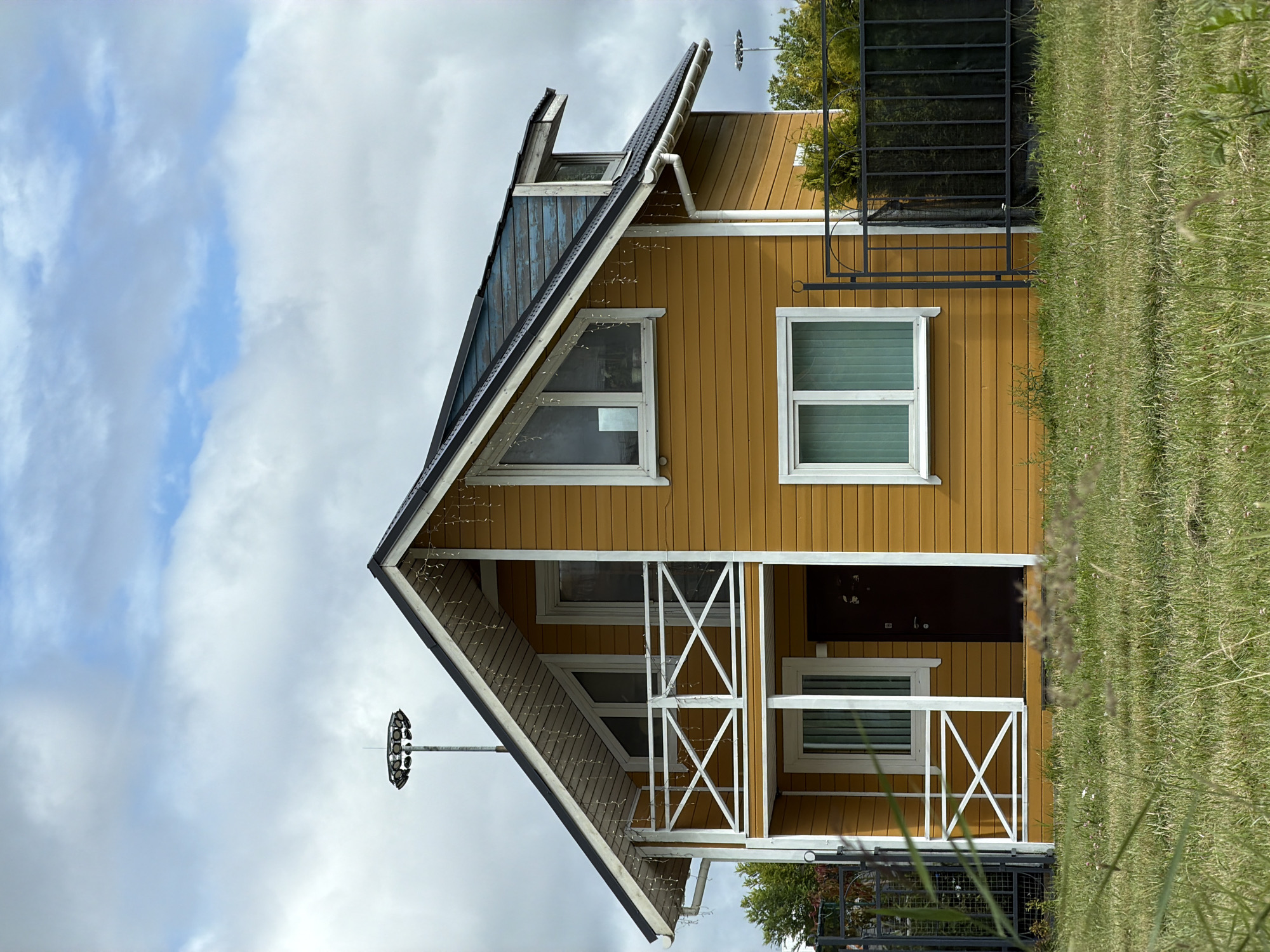 A two-story wooden house with yellow siding and a dark roof is depicted in the image. The house has a prominent balcony on the second floor with white railings. Several windows are visible, including triangular ones on the upper level and rectangular ones on both floors. White string lights are draped along the eaves of the roof and the balcony railing. In the background, a tall pole with a circular light fixture is visible. The house is surrounded by a black metal fence and sits on a grassy area. The sky is overcast with patches of blue. The scene appears to be a residential property in Pargolovo, Russia.
