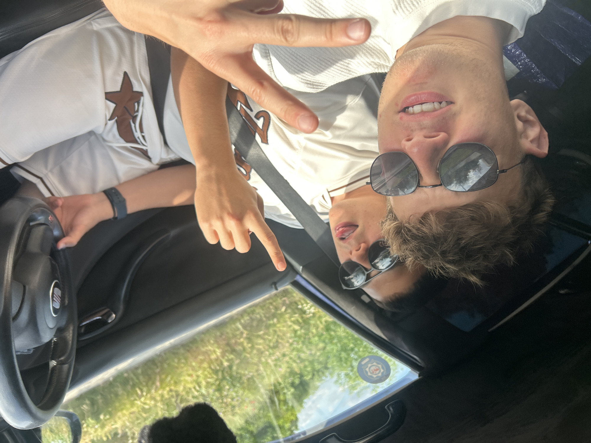 The image shows a young man inside a car. He is wearing sunglasses and a white baseball jersey with brown trim and a star emblem. He's making a "peace" sign with one hand and pointing with the other. The car's steering wheel is visible, along with part of the dashboard and the view through the windshield, which shows greenery. The image is oriented upside down. The location in the city is not specified.