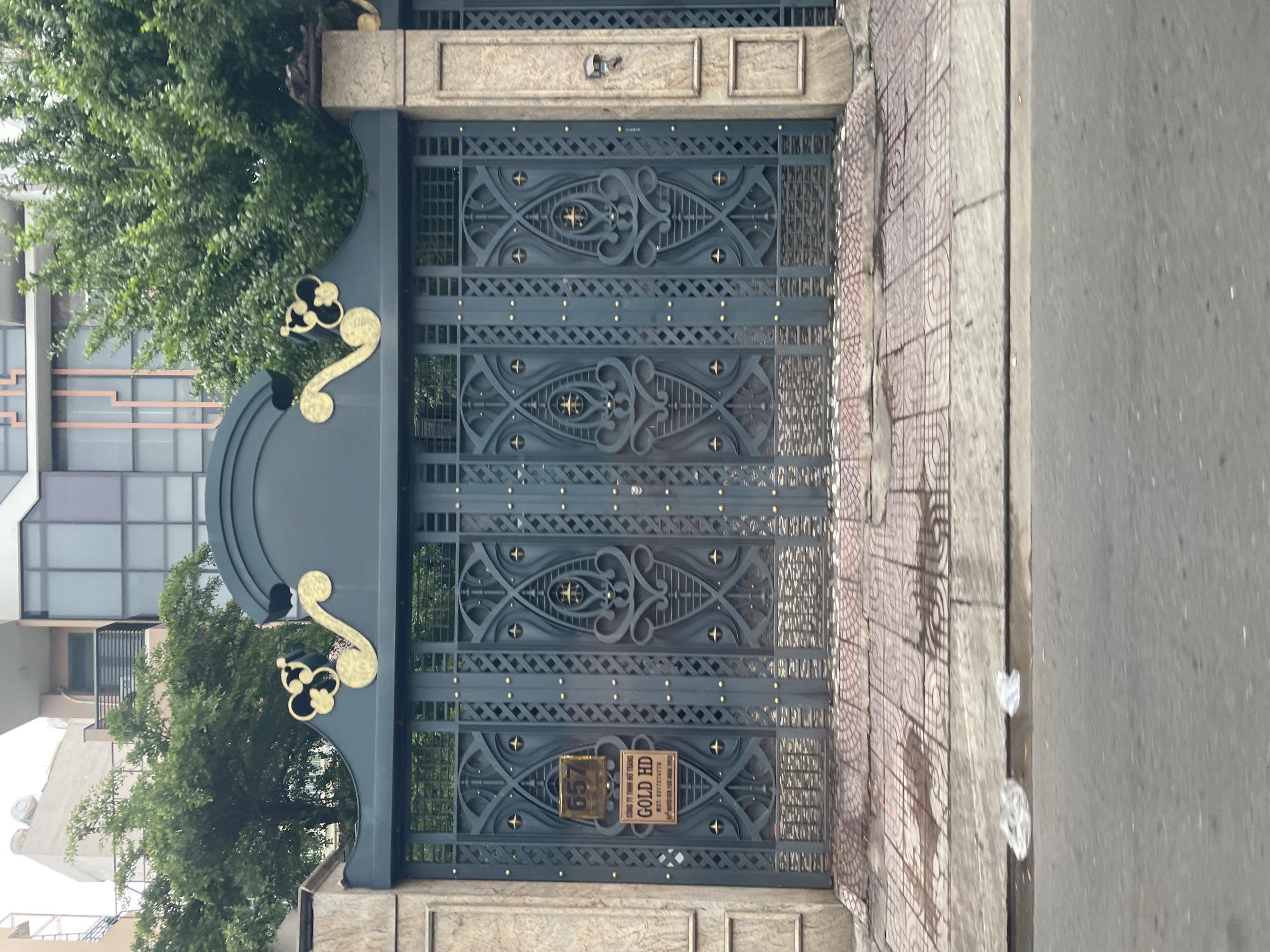 The image shows a large, ornate dark gray metal gate with gold decorative elements and a gold plaque reading "657" on the left panel. Below this is another gold plaque with text in Vietnamese and "GOLD HD" in English. The gate is set within stone pillars and is on a sidewalk paved with red and gray tiles. In the background, there are green trees and modern buildings. The street in front of the sidewalk is asphalt. There are two pieces of crumpled white plastic on the edge of the sidewalk near the street. The scene is located in Thủ Đức, Vietnam.