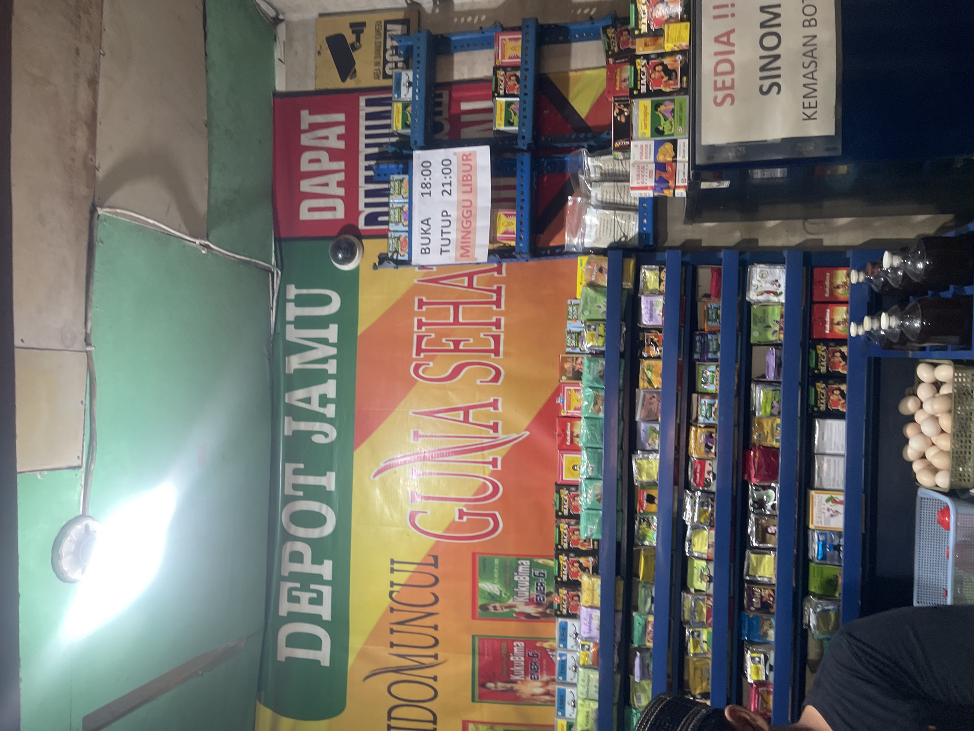 This image captures a storefront in Kebonagung, Indonesia, likely a small shop selling traditional Indonesian herbal remedies (jamu). The scene is oriented vertically, with most of the signage running from bottom to top.
The main focus is on a large, colorful banner that reads "DEPOT JAMU INDOMUNCUL GUNA SEHA," suggesting the name of the establishment and that it offers beneficial remedies. To the left of this banner, a red sign states "DAPAT," which translates to "get" or "obtain," likely indicating that certain products are available. A surveillance camera is visible mounted on the wall near the signage.
The backdrop features shelves stocked with numerous small, packaged items, likely the herbal products themselves. Some of these packages have visible text and imagery, though they are too small to decipher clearly. A white sign is posted, providing operating hours: "BUKA 18:00" (Open 18:00) and "TUTUP 21:00" (Close 21:00), with "MINGGU LIBUR" (Sunday Closed) indicating their weekly schedule.
To the right of the jamu shelves, a dark blue cabinet or structure is visible, with more shelves containing what appear to be bottles. Some round, light-colored objects, possibly eggs, are contained within a basket on top of this structure.
A portion of a person wearing a black shirt and a peci (a traditional cap) is visible in the lower right corner, suggesting a customer or the proprietor. The lighting appears to be artificial, with a bright light source illuminating the left side of the image, causing some glare on the green wall. The overall impression is of a modest, functional shop selling traditional Indonesian goods.