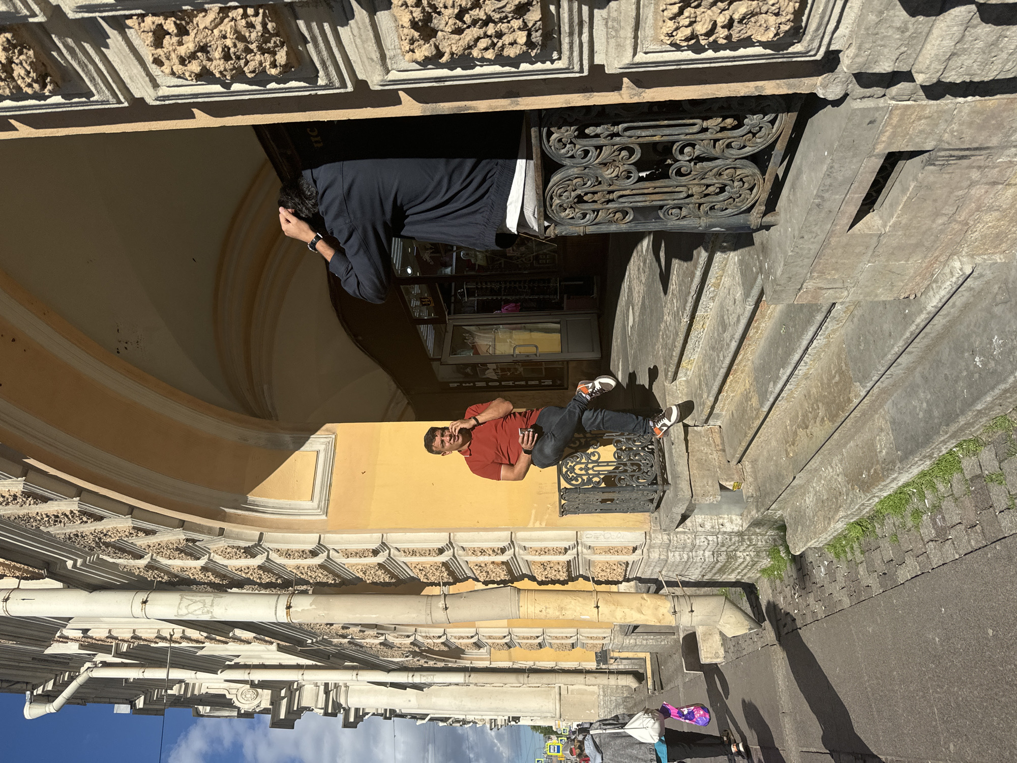 Two men are outside a building in Saint Petersburg, Russia. The building has ornate architectural details and a bright yellow facade.
In the foreground, a man in a red polo shirt and jeans is sitting on a decorative metal railing. He is holding a phone in his right hand and appears to be talking on it. He has short dark hair and is wearing sneakers.
Behind him, and further inside an arched entryway, another man is standing and looking down, with his hands near his face as if contemplating something. He is wearing a dark jacket and light-colored pants. The entryway leads to a shop with a display window.
The scene suggests a sunny day, with strong shadows cast by the building and the people. The overall atmosphere appears to be casual, with one man engaged in a phone call and the other in a thoughtful pose. The architectural style of the building, with its decorative stonework and the presence of iron railings, is characteristic of historical European cities like Saint Petersburg.