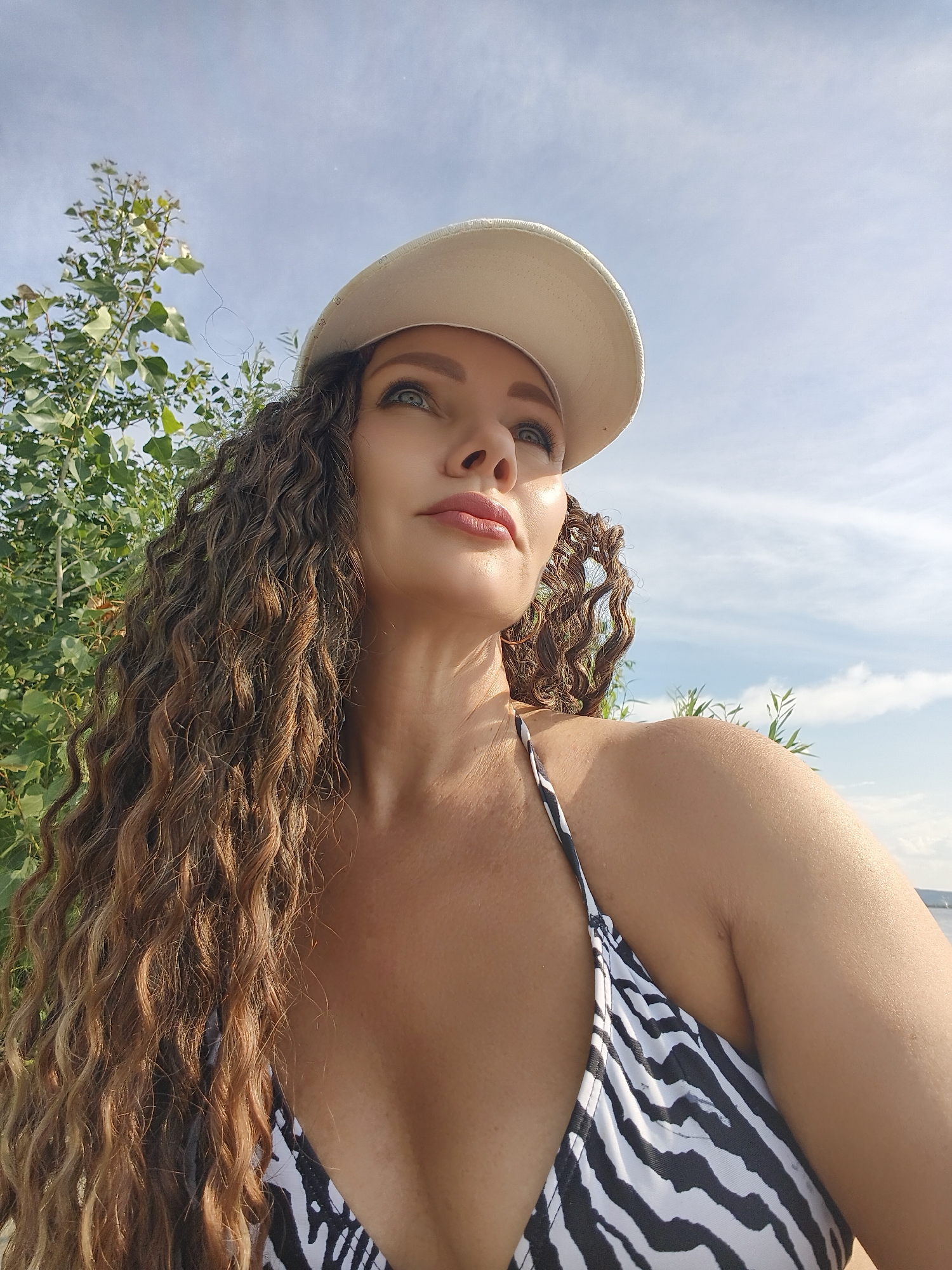 A low-angle, medium shot captures a woman from the chest up, wearing a white baseball cap and a black and white zebra-print bikini top. She has long, curly brown hair, and her head is tilted upwards, looking towards the sky. Her skin is tanned, and there is sunlight reflecting on her forehead and shoulder. She is outdoors, with green foliage visible behind her on the left, and a bright blue sky with scattered clouds occupying the upper portion of the frame. The background is blurred, suggesting a shallow depth of field. The location is unknown.