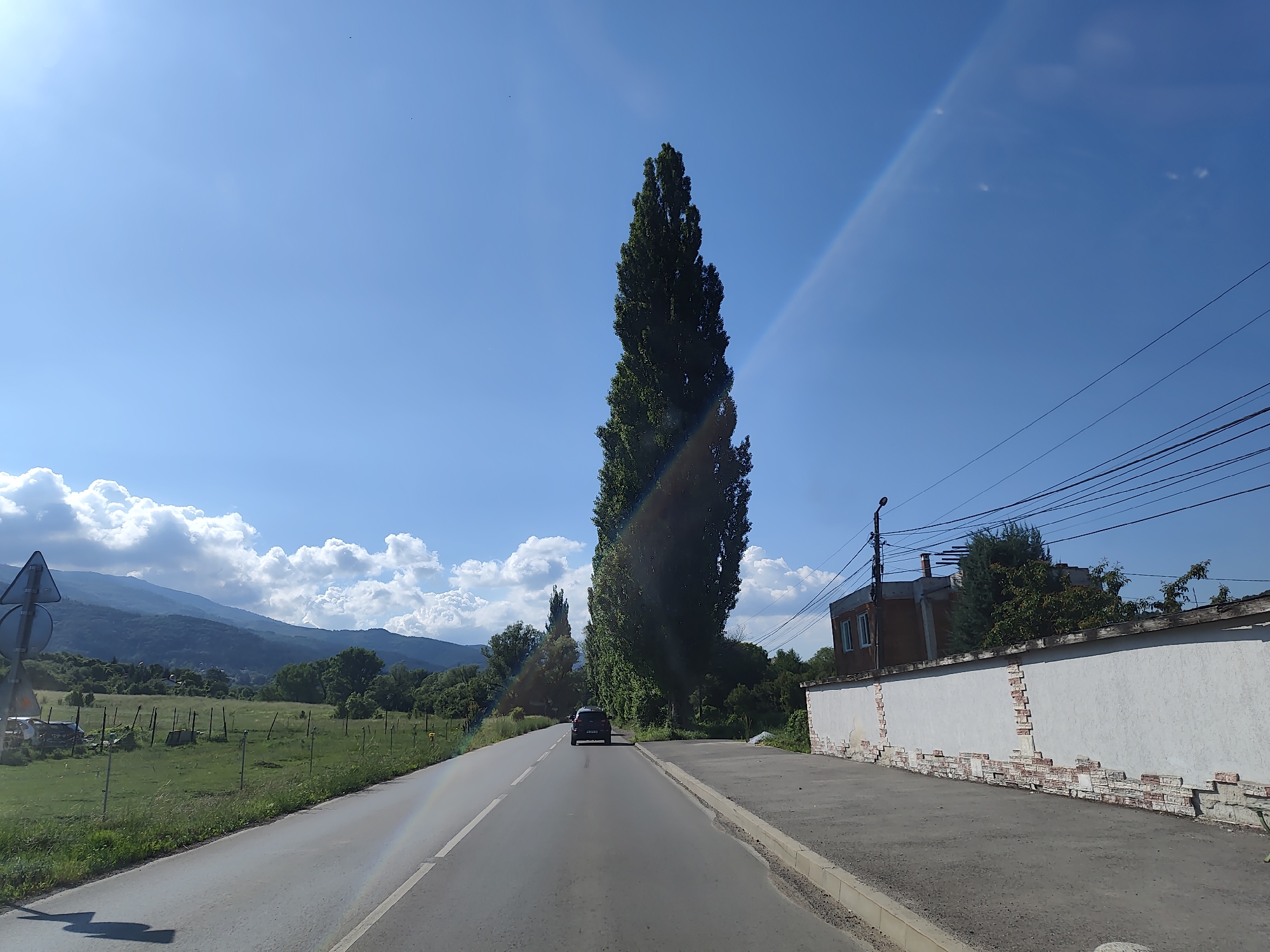 The image depicts a road in the city of Dragichevo, Bulgaria, on a clear, sunny day. A dark-colored car is driving on the asphalt road, which has white lane markings. To the left of the road, there is a grassy field with scattered wooden posts and a traffic signpost. In the background, rolling hills covered in trees and vegetation are visible under a blue sky with scattered white clouds. A tall, slender tree stands prominently near the center of the image. To the right of the road, there is a white wall with decorative brickwork at its base, and a building with windows and a sloped roof behind it. Overhead power lines are strung across the upper right portion of the scene. The overall impression is of a rural or suburban road with natural and man-made elements.