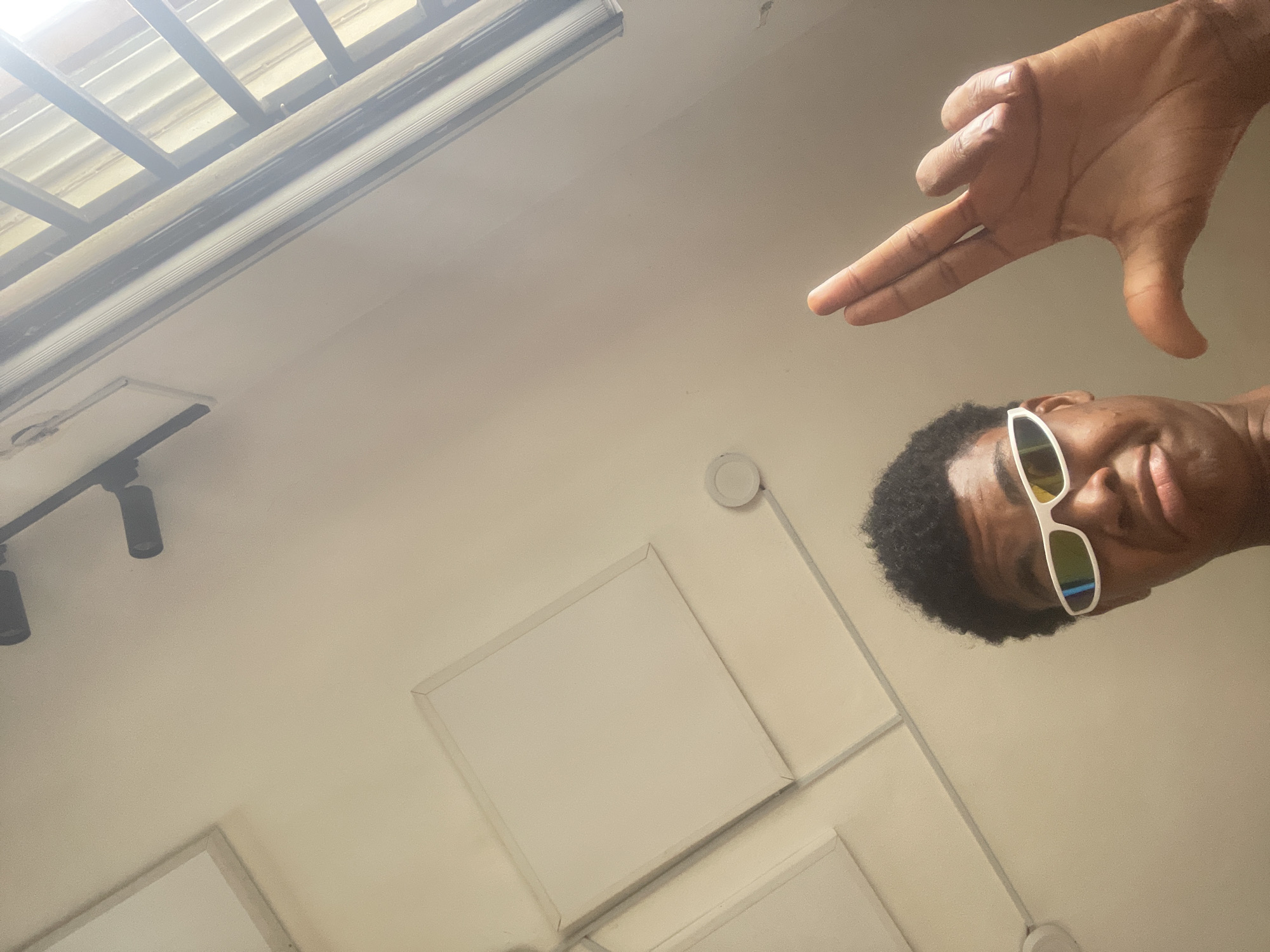 An overhead, low-angle, full shot captures a young Black person wearing bright white, reflective wraparound sunglasses, looking directly at the camera. Their expression is a slight pout, with lips pursed. The person is giving a peace sign with their right hand, holding up two fingers in a V-shape, with their thumb extended. Their afro hair is short and curly. The background consists of a pale, textured ceiling with recessed square panels and some ceiling-mounted lights. The scene appears to be indoors.