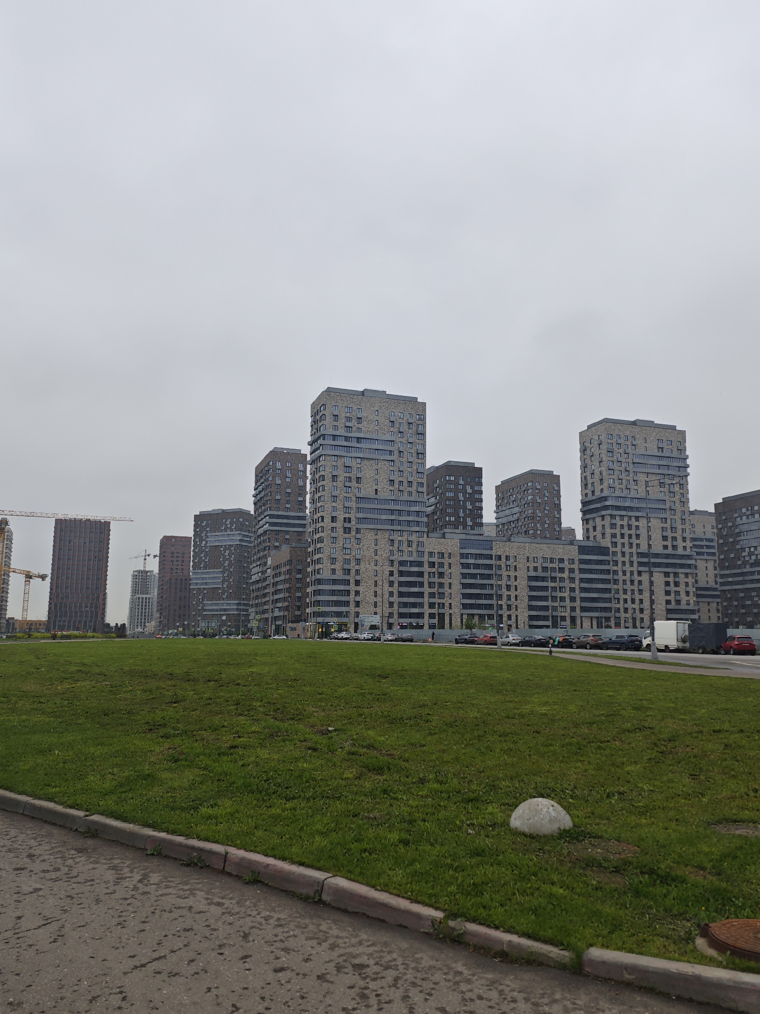 The photograph depicts an urban landscape on an overcast day in Khimki, Russia. In the background, a series of modern high-rise apartment buildings of varying heights dominate the skyline. The buildings are constructed with facades featuring a mix of brick and glass elements. Construction cranes are visible in the distance, suggesting ongoing development in the area.
A broad expanse of green grass occupies the foreground. A concrete curb separates the grass from an asphalt surface in the lower part of the frame. A spherical concrete bollard is visible on the grassy area near the curb.
Along the road in front of the buildings, multiple cars are parked or in transit. Pedestrians are visible walking along the side of the road.