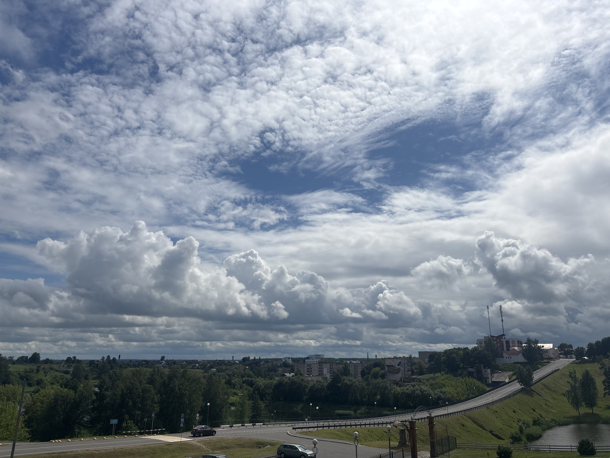 An outdoor, wide-angle, high-angle photograph shows a view of the city of Капыльскі сельскі Савет, Belarus, under a dramatic sky filled with large, fluffy white and grey clouds. In the foreground, a grey asphalt road curves to the right, bordered by a grassy slope. Several cars are visible on the road, including a dark sedan in the left lane and a silver sedan in the right lane. A pedestrian crossing is marked on the road.
Further into the scene, a bridge with a white railing spans over a body of water. Beyond the bridge, a town unfolds with numerous trees, buildings of various sizes and architectural styles, and some distant industrial structures or towers. A prominent building with red accents and communication towers sits on a hill to the right. The overall impression is one of a partially overcast day with a blend of natural landscape and urban development.