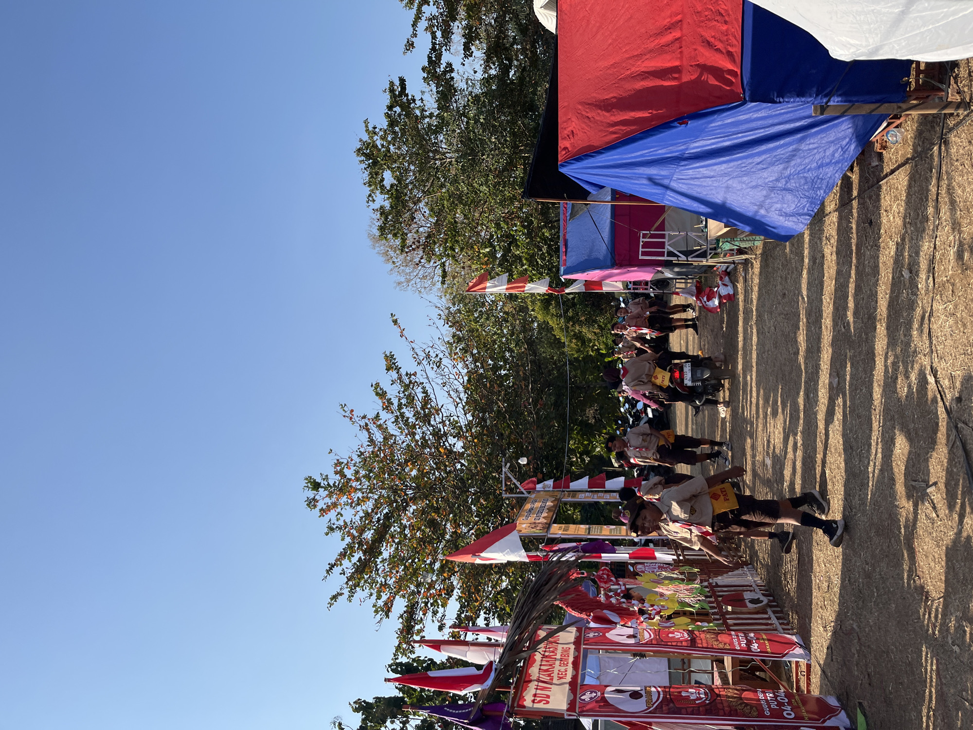 In Gembong, Indonesia, a group of people, primarily young scouts in uniform, are walking along a dirt path lined with tents and banners. The event appears to be a scout gathering or camp. The scouts are wearing khaki shirts and shorts, some with badges and neckerchiefs. Several individuals are walking towards the camera, while others are seen in the background.
To the left of the path, there are several red and white banners with text and logos, indicating a local school or scout troop, possibly "SD N 02 KARKASIH" and "KEC. GEMBONG." Flags, including Indonesian flags, are also visible.
On the right side of the path, there are several tents, including a large red tent and a blue tent, suggesting a camping or event setup. A motorcycle is parked behind some of the scouts in the middle ground. The environment is outdoors, with trees and a clear blue sky. The ground is covered in dry grass and dirt, and shadows from the tents and trees are cast on the path. The overall scene suggests a daytime event in a rural or semi-rural setting.