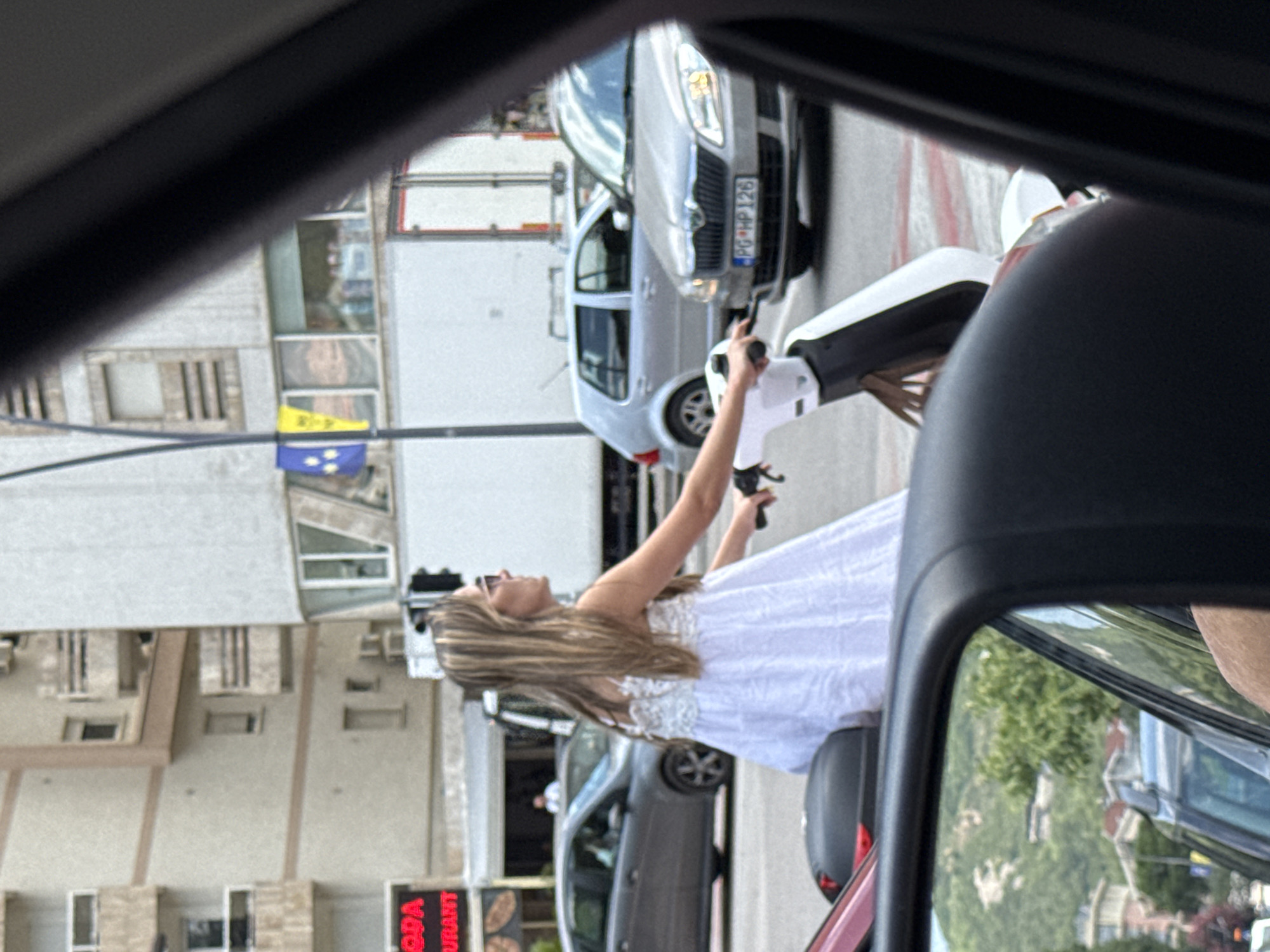 A woman with blonde hair, wearing sunglasses and a white dress, is riding a white electric scooter. She is on a city street, with buildings and cars visible in the background. The license plate of a silver Skoda car in front of her reads "PG HP 126". The scene appears to be in Budva, Montenegro, given the context of the plate's format and the typical urban scenery. A portion of the photographer's vehicle, including the side mirror, is in the foreground, suggesting the image was taken from a car.