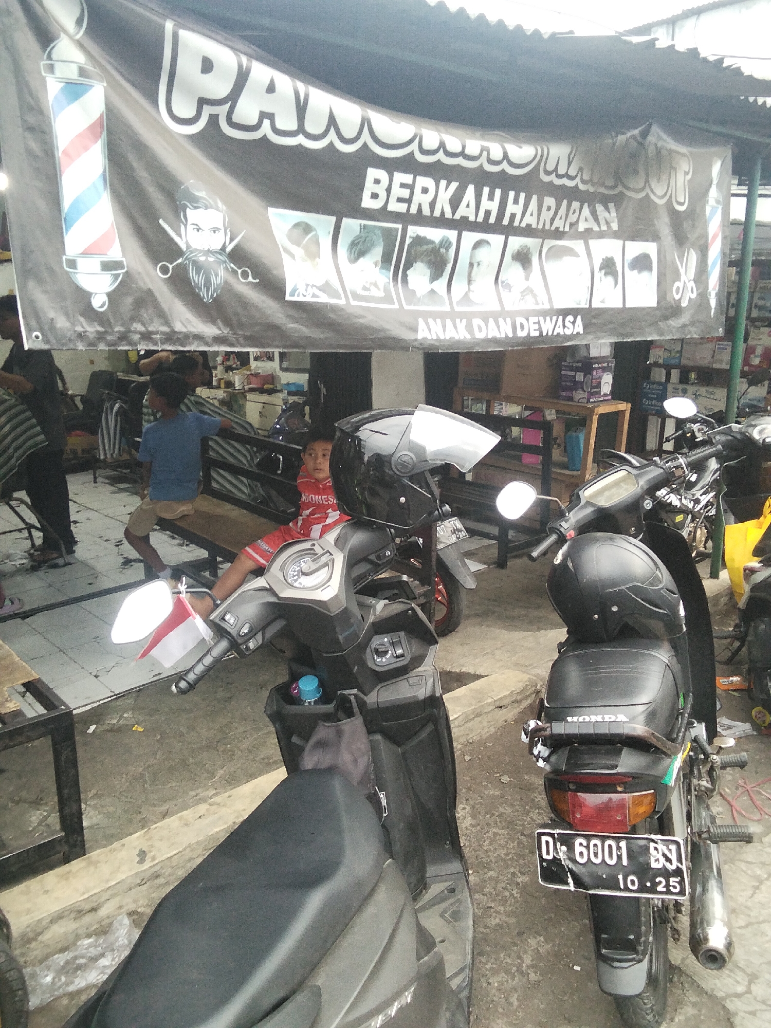 A banner with a barber pole and images of hairstyles hangs above a street scene. In the foreground, two motorcycles are parked. A young boy in a red and white shirt is sitting on one of the motorcycles, with his hand on the handlebar. A red, white, and blue Indonesian flag is attached to the handlebar. In the background, people are visible inside what appears to be a barbershop. Shelving units with various products are also present in the background. The ground is paved with concrete.