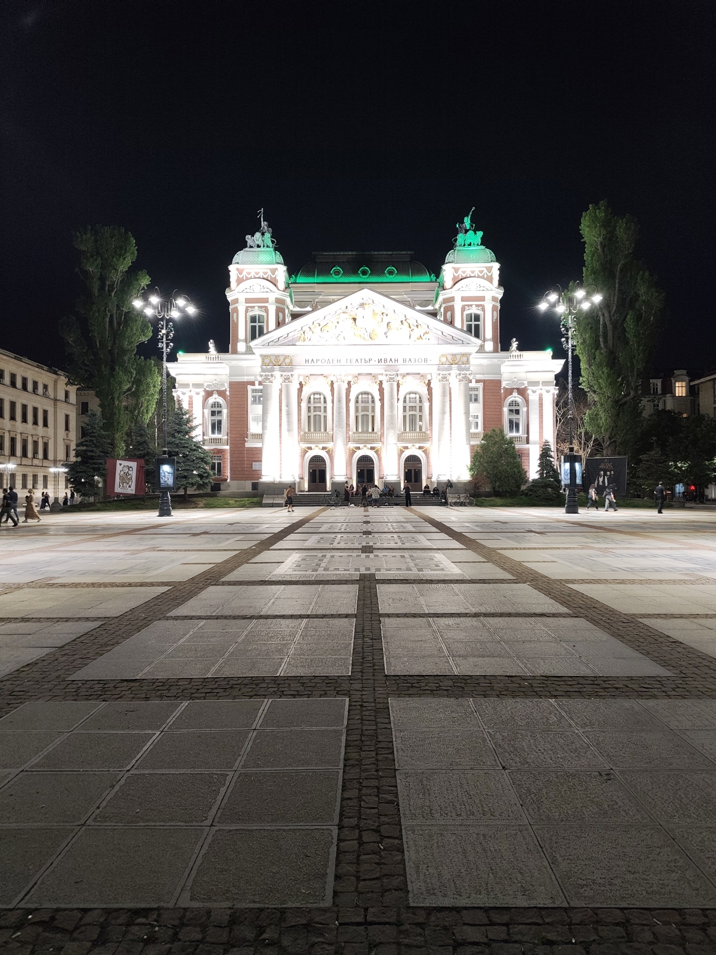 The image depicts the Ivan Vazov National Theatre in Sofia, Bulgaria, at night. The theatre is illuminated with bright white light, showcasing its architectural details. The square in front of the theatre is paved with large square stones separated by darker cobblestones. Tall trees frame the building on either side, with streetlights visible amidst the trees. Several people are walking around the square, some near the building and others in the distance. Movie posters are displayed on the left and right side of the square. The sky is dark, suggesting it is late evening or night.