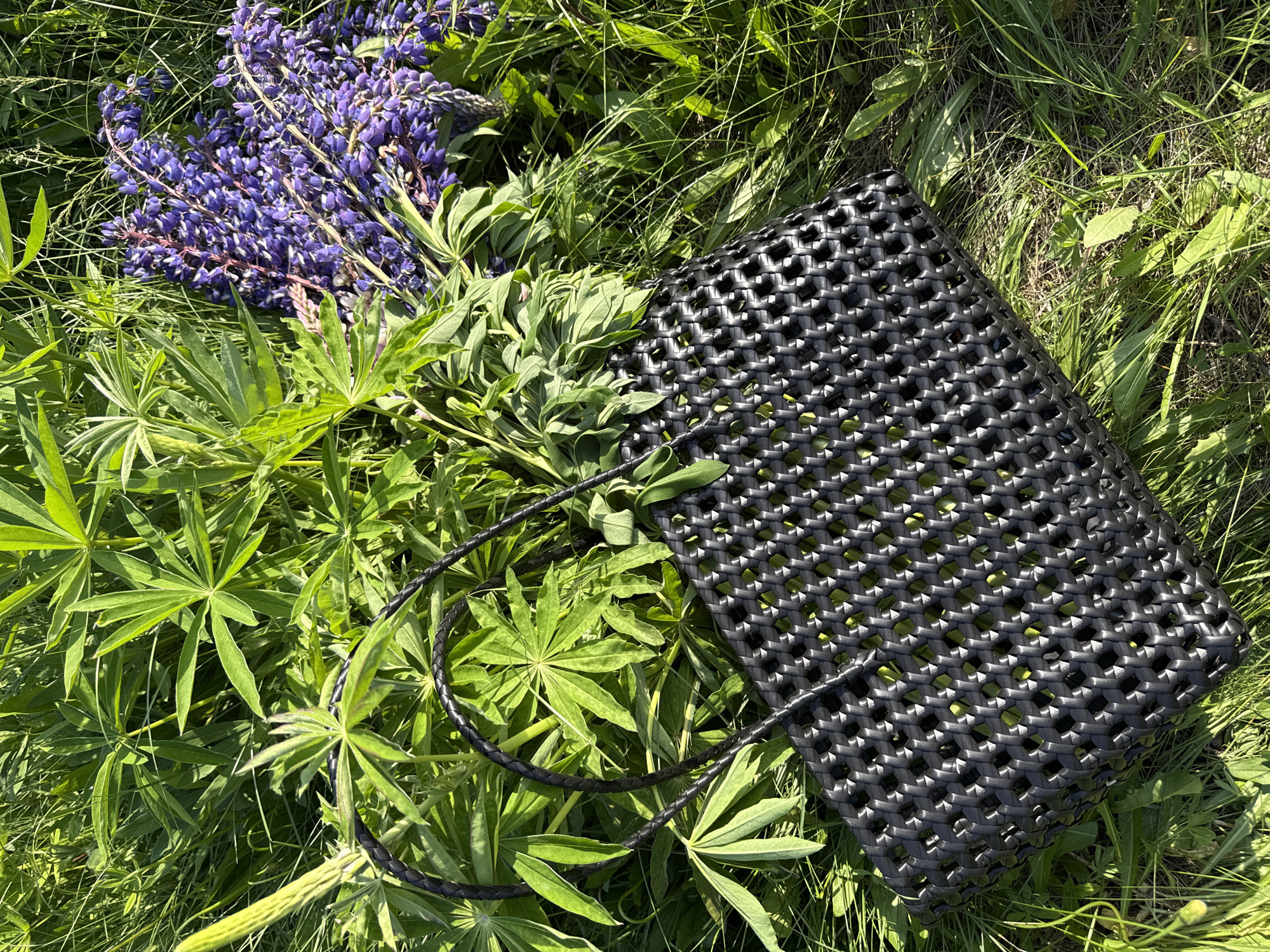 In the city of Chaikovsky, Russia, a black woven tote bag and a bundle of purple lupine flowers are situated amidst dense green vegetation. The black bag, positioned in the lower-left to mid-right foreground, features a tightly interlaced pattern of flat strips, forming a grid-like structure. It is equipped with two braided black handles. To the upper right of the bag, a cluster of freshly cut purple lupine flowers, characterized by their tall, dense spires of individual blossoms and visible pinkish stems, rests horizontally. The surrounding environment is dominated by vibrant green foliage, including numerous large, palmate leaves typical of lupine plants, along with various tall grasses and other ground cover. The scene is brightly illuminated by strong, direct sunlight, casting distinct shadows and highlighting the textures of the plants and the bag. No people or animals are visible.