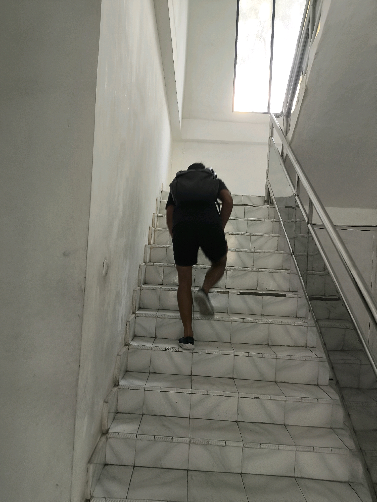 An individual wearing a dark backpack and black shorts walks up a tiled staircase. The staircase is bordered by a white wall on the left and a metal railing on the right. Natural light streams in from a narrow, vertical window at the top of the staircase. The scene is indoors, likely in a residential or public building.