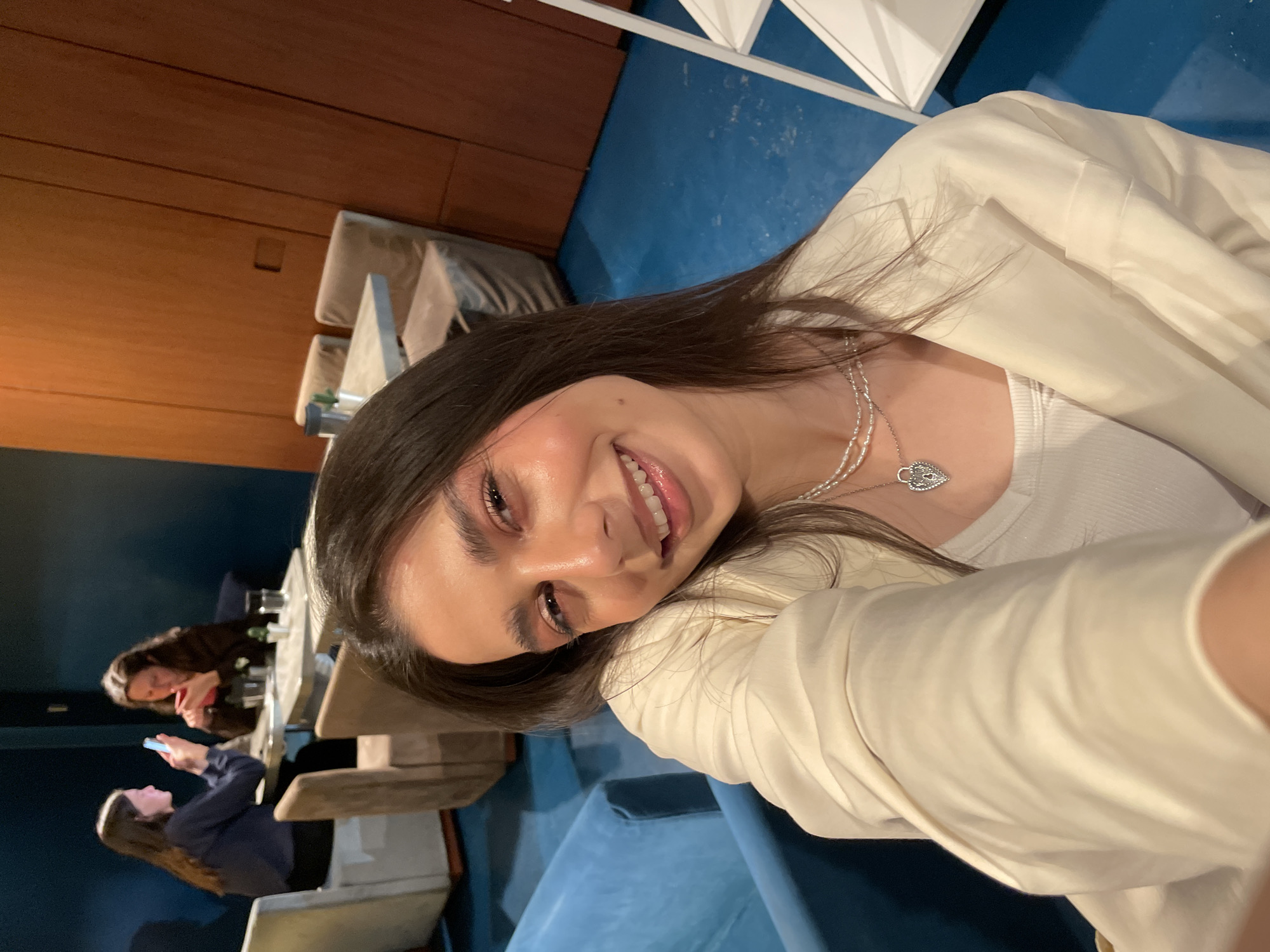 A smiling woman takes a selfie in an indoor setting. The background includes tables with people, one using a phone. The interior features a blue floor, wooden paneling, and modern chairs. The woman wears a white jacket, a white top, and a necklace. The location is likely a restaurant or similar establishment in Milan, Italy.