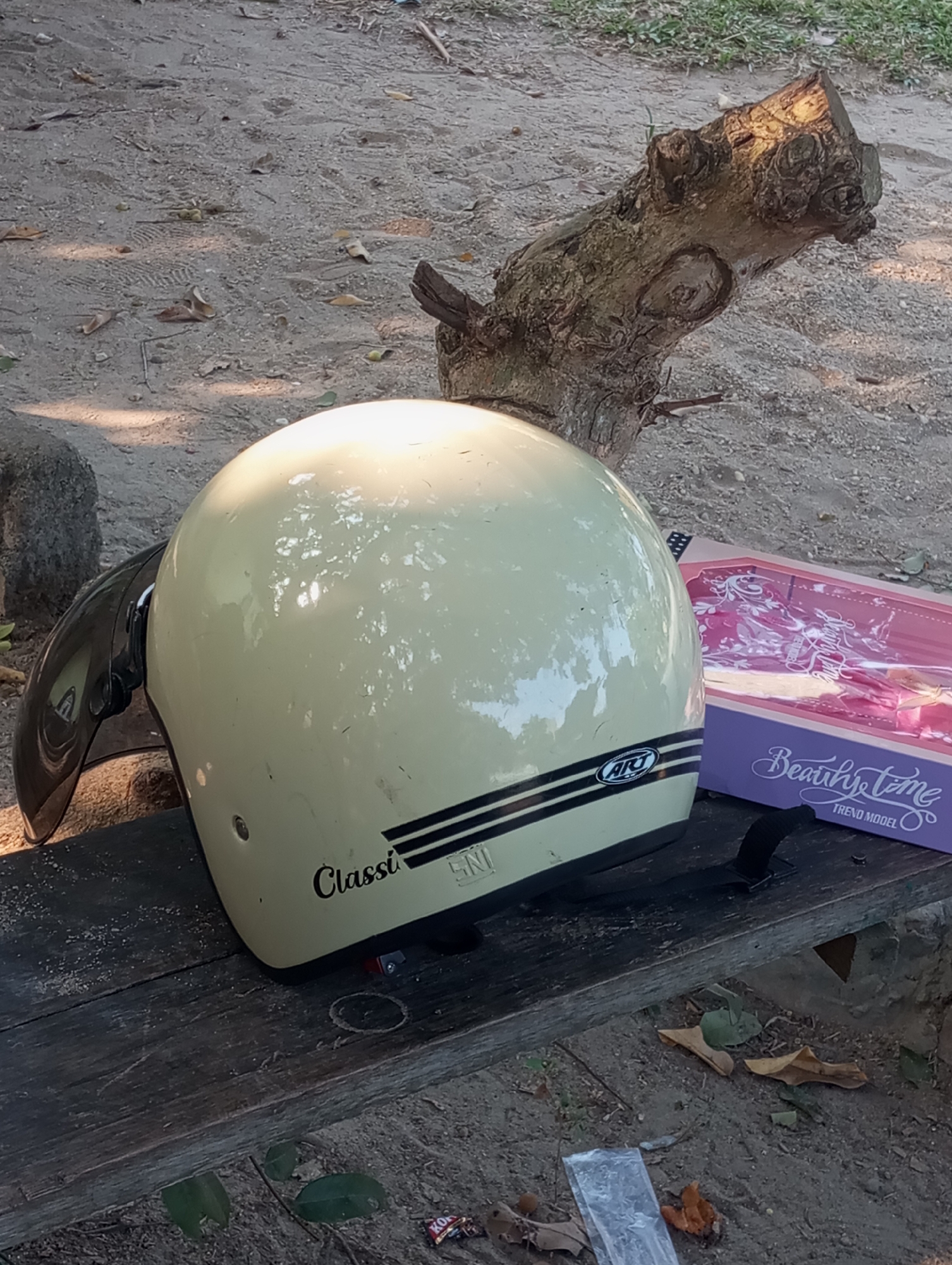 A cream-colored motorcycle helmet with black and white stripes and a black visor rests on a weathered wooden bench. To the right of the helmet is a purple and pink box with "Beauty Time" written on it, suggesting it contains a doll. In the background, a thick, gnarled tree branch lies horizontally on sandy ground. The scene appears to be outdoors, possibly in a park or yard, with dappled sunlight filtering through unseen foliage. The presence of a log and sandy ground suggests a natural or semi-natural environment within Pematang Reba, Indonesia.