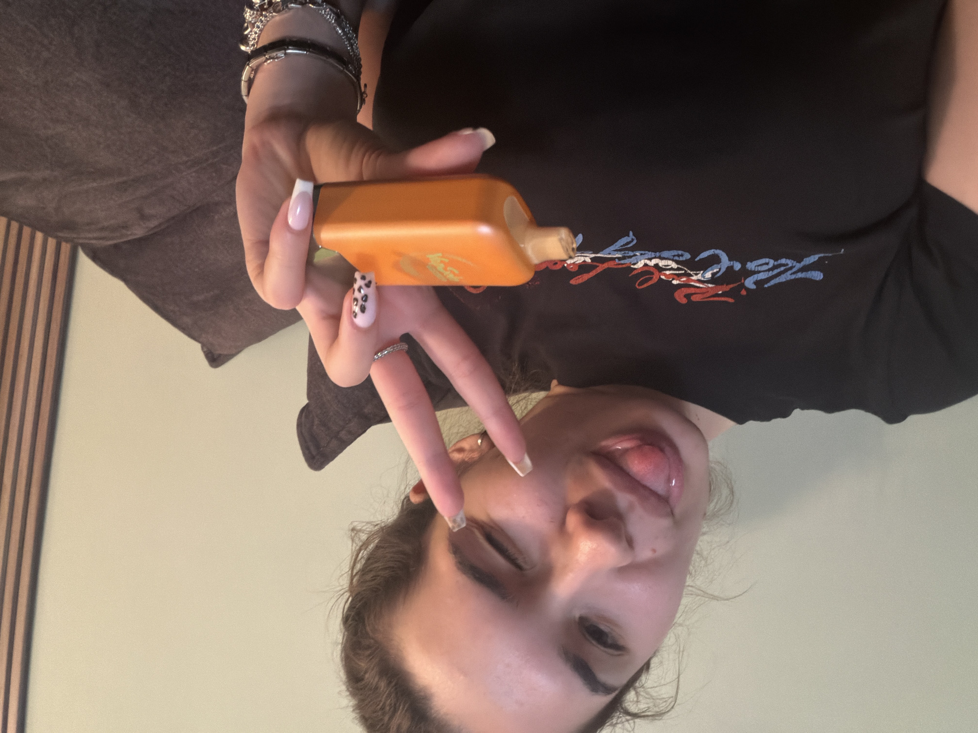 A selfie of a person with light skin is shown. They are wearing a black t-shirt with a red, white, and blue design on it. The person is holding an orange bottle with a nozzle in their hand, flashing a peace sign. They have jewelry, including multiple bracelets and a ring, on their hand. Their fingernails are painted, one with a pink base and black spots. The background consists of a brown couch and a light green wall with vertical wood panels. There is no evidence to suggest that this picture was taken in Kavarna, Bulgaria.