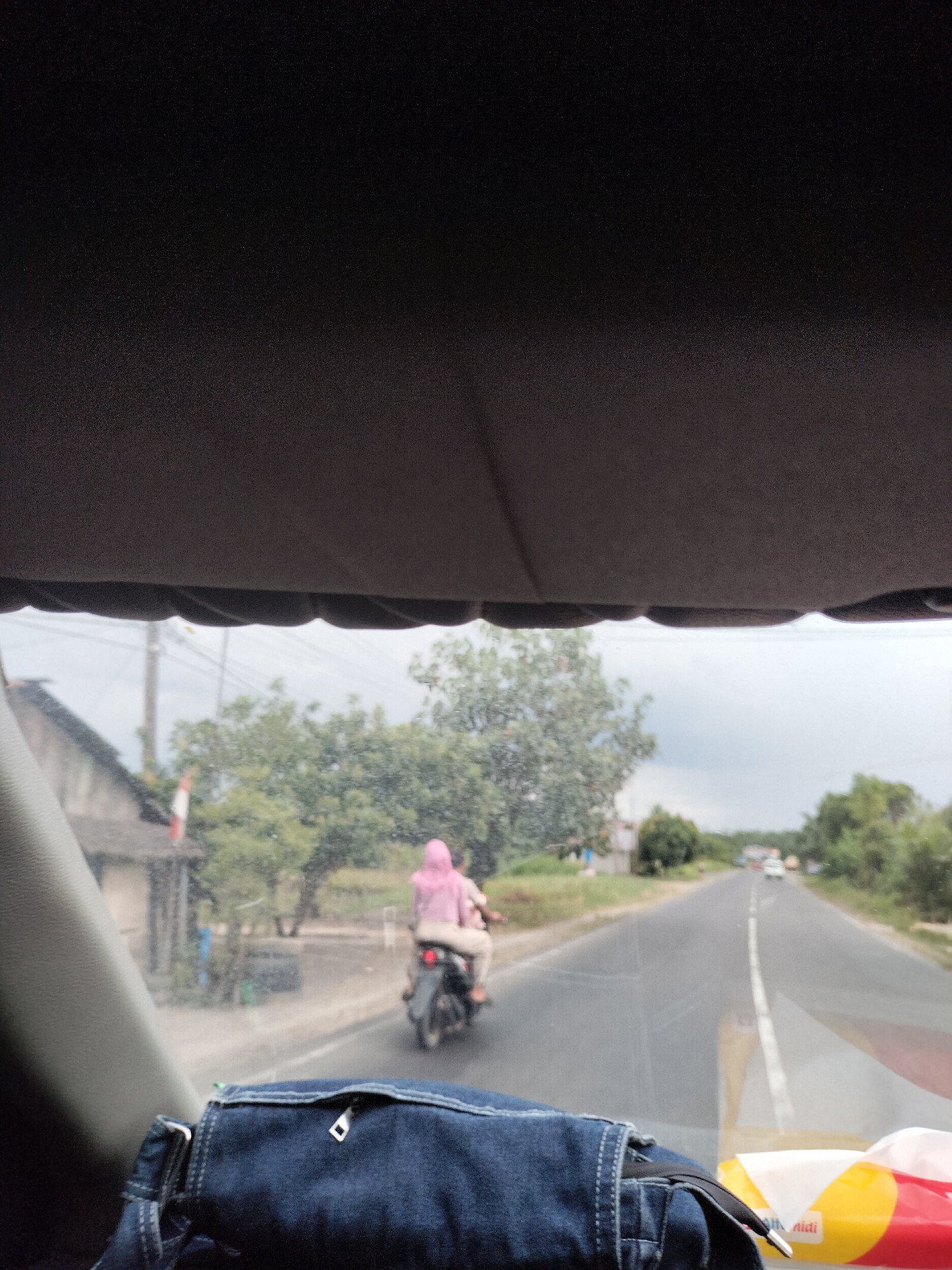 The image shows a view from inside a vehicle looking out onto a road in Bukit Selamat, Indonesia. In the foreground, a blue denim bag with a zipper is visible. Beyond the bag, through the windshield, two people are riding a motorcycle. The person in the back is wearing a pink hijab. They are traveling on a paved road with a white line dividing the lanes. To the left of the road, there is a building with a dark roof and a small flag. Trees and vegetation line the sides of the road. The sky is overcast.