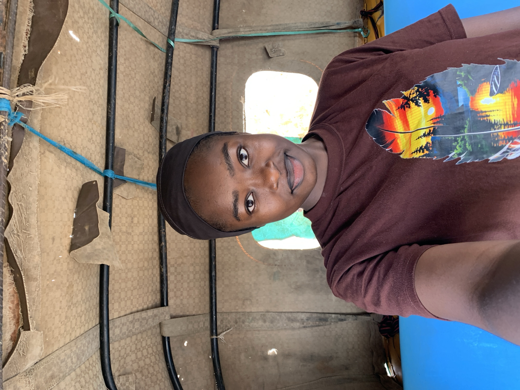 A young Black person wearing a brown t-shirt with a feather graphic and a black headscarf is looking directly at the camera, seemingly inside a vehicle with a textured interior. The vehicle has black metal supports, and some of the interior material appears worn or torn. Through a window, one can see a blurred glimpse of the outside. The image was taken in Rafin Par, Nigeria.