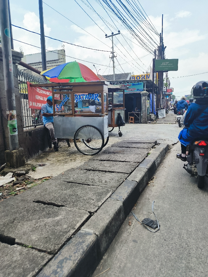 A food cart, labeled "Special Batagor", sits parked on the sidewalk in Ngamprah, Indonesia. The cart is made of wood and metal with a white canopy and a large, black tire on the left side. There is a person standing next to the cart, but their face is obscured from view. The cart is parked on the sidewalk, which is made of grey concrete and bordered by a black curb. There are a number of power lines overhead, with one pole visible in the background, to the left. The sidewalk is relatively empty, save for a few pieces of litter. The person on the food cart appears to be interacting with a person who is riding a scooter. The scooter is parked in the middle of the image, and the rider is wearing a blue jacket. The image was likely captured on a clear day.