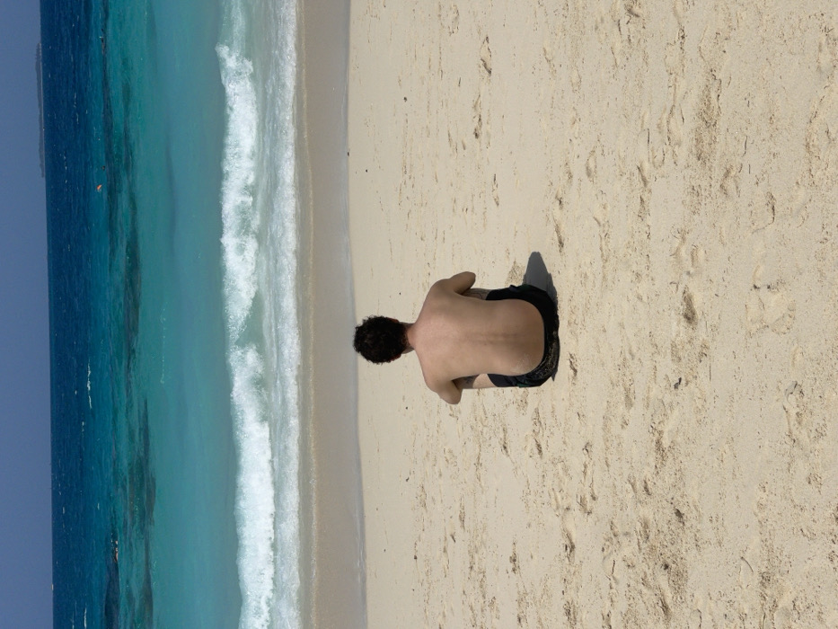 An individual, presumed male, is seated alone on a light-colored sandy beach in Zanzibar, Tanzania. The subject is observed from behind, shirtless, with dark, curly hair, wearing dark-colored swim trunks, and is positioned with knees bent, facing the ocean. The immediate foreground consists of sandy beach with visible indentations. Beyond the sand, a line of white breaking waves transitions into distinct zones of clear turquoise and deeper blue ocean water. A small, orange, floating object is visible in the mid-distance water. A faint, dark landmass, consistent with an island, is discernible on the distant horizon to the right, beneath a clear blue sky.