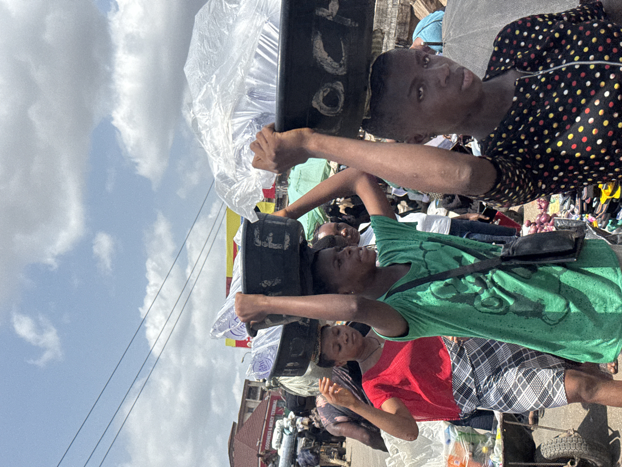 This image captures a vibrant, bustling outdoor market or street scene in Onitsha, Nigeria, during daylight hours under a cloudy sky. The perspective is slightly elevated, looking down into a crowded area.
The primary subjects are several individuals, mostly young people or adults, engaged in market activities. In the foreground and center, a young woman in a black and multi-colored polka dot long-sleeved top looks directly towards the viewer. She is carrying a black container or tray on her head, which appears to hold items wrapped in clear plastic bags. Her right arm is raised, possibly to steady the load.
Behind her, another person in a bright green t-shirt with visible sweat stains on the back is also carrying a black container on their head, holding it with both hands. They are also wearing a black shoulder bag. Further back, another person in a red t-shirt and plaid shorts is visible, with a hand raised. Many other individuals are scattered throughout the background, some appearing to be vendors with stalls or goods, while others navigate the crowded space. One crop reveals a pile of what looks like onions or similar vegetables on the ground near a tire.
The setting is an active street or market area with buildings in the background. One building clearly displays a sign that reads "GOODNESS & MERCY TERMINAL." Utility poles and power lines crisscross the upper left portion of the sky. The overall impression is one of a busy commercial environment where goods are transported and sold, reflecting daily life in an urban Nigerian market. The writing "OCHO" is faintly visible in white on one of the black containers carried on a head.