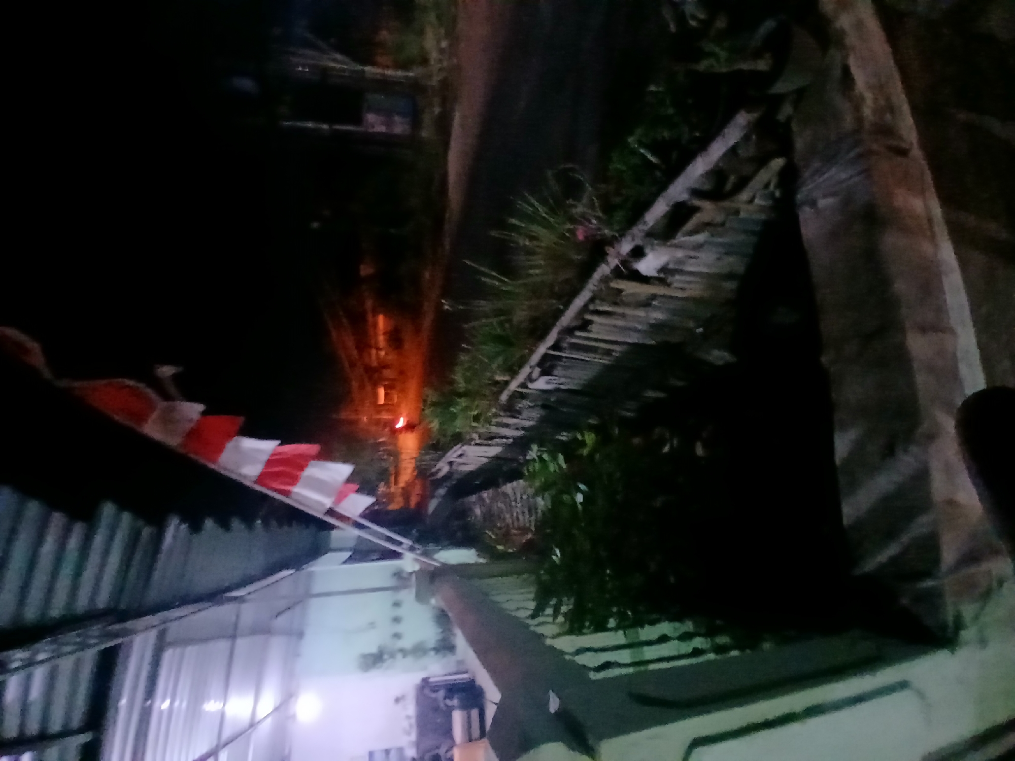 A nighttime view from a dimly lit porch, looking out towards a road and vegetation. A string of Indonesian flags hangs from the roofline. A narrow ditch runs alongside the road, bordered by a wooden structure and dense greenery. The road itself is dark, with a faint orange glow visible in the distance, possibly from a vehicle's headlights or a distant light source. The environment appears to be a residential area in Kademangan Kulon, Indonesia.