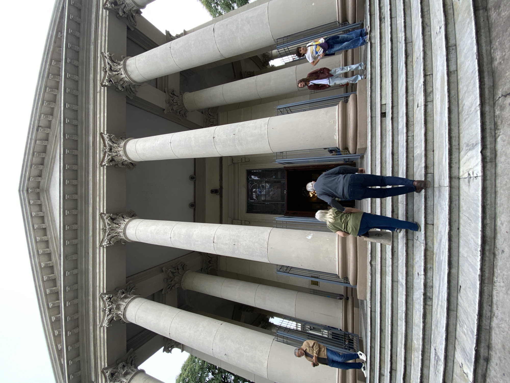 The image depicts the entrance to the Metropolitan Cathedral of Buenos Aires in Argentina. The cathedral's neoclassical facade, characterized by large columns and intricate architectural details, dominates the frame. Several people are ascending the wide staircase leading to the entrance. The steps appear to be made of marble. Some individuals are closer to the door, while others are further down the stairs. The lighting is somewhat diffused, suggesting an overcast day.