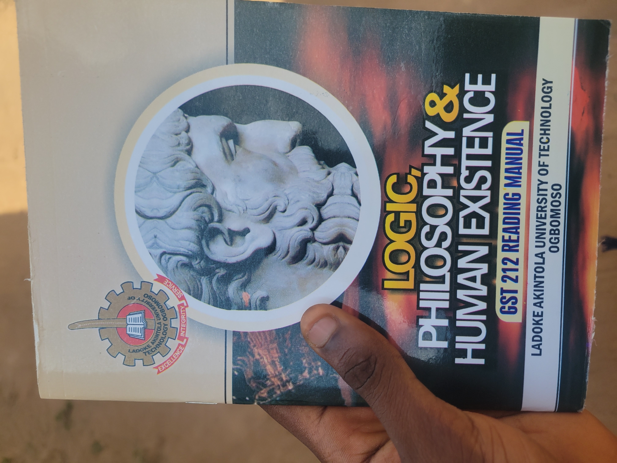 The image is a close-up of a book cover held by a person's hand. The book is titled "LOGIC, PHILOSOPHY & HUMAN EXISTENCE" and is a "GST 212 READING MANUAL". It is published by Ladoke Akintola University of Technology, Ogbomosho. The cover features a circular image of a classical sculpture of a face, possibly of a Greek or Roman deity, with intricate details of hair and beard. The background of the book cover has a fiery, abstract design. On the left side of the book cover, there is a circular emblem with text that appears to be the university's logo, containing the words "SERVICE", "INTEGRITY", "EXCELLENCE", "LADOKE AKINTOLA UNIVERSITY OF TECHNOLOGY", and "OGBOMOSO". The image appears to be taken outdoors, with the lighting suggesting it could be daytime, though no specific time is discernible. The focus is on the book and the hand holding it, with the background slightly blurred, providing no further contextual cues about the location.